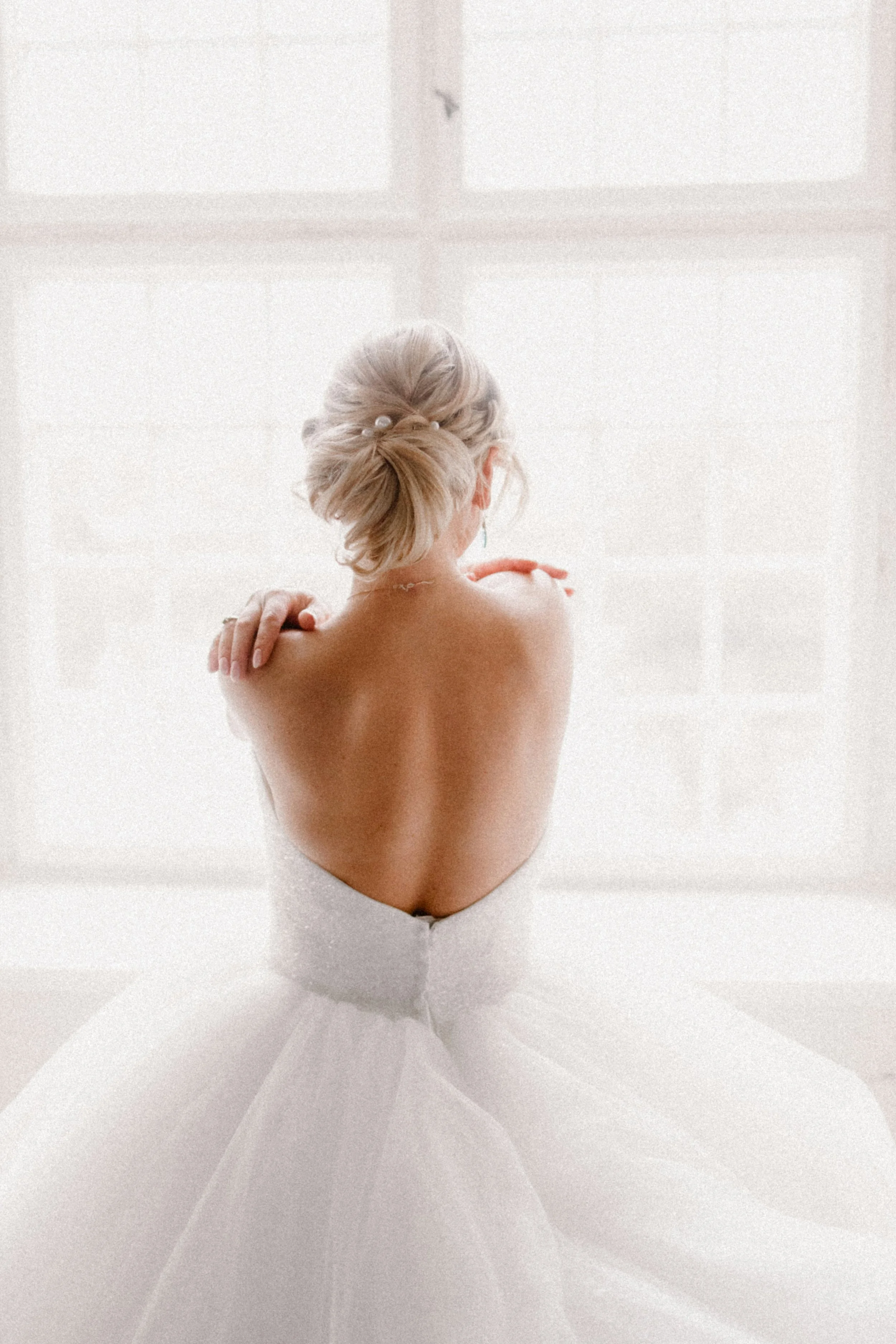 A woman in a white wedding dress, turned away from the camera, standing in front of a large window with sheer curtains.