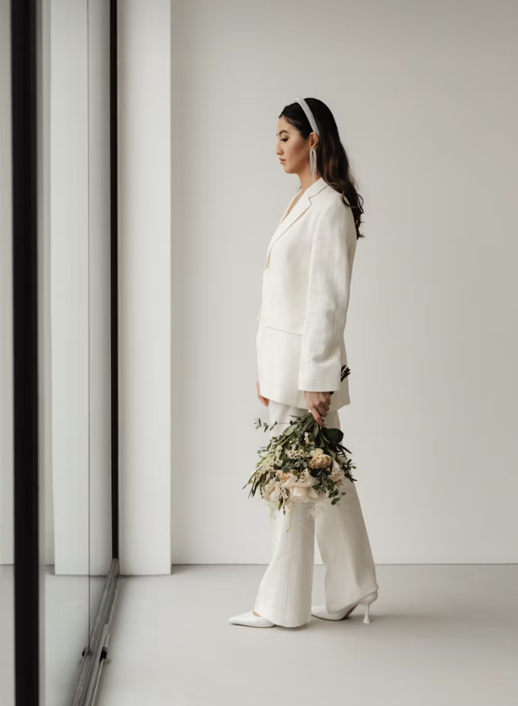 A woman in a white suit with wide-leg pants and a blazer, holding a bouquet of flowers, standing in a minimalistic room with white walls and a mirror.