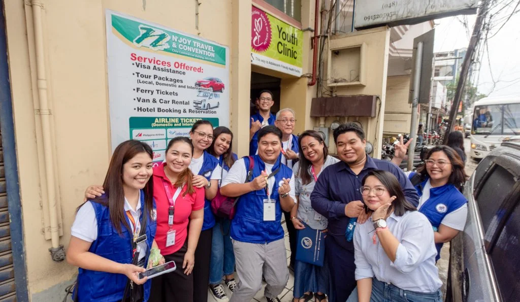 Group of people smiling and posing for a photo outside a building with a sign for a travel and youth clinic, some wearing blue vests and others in casual clothing.