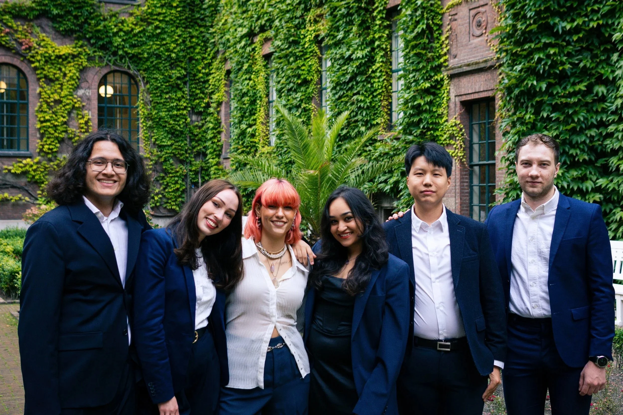 Six young adults in business attire posing together outdoors in front of an ivy-covered building.