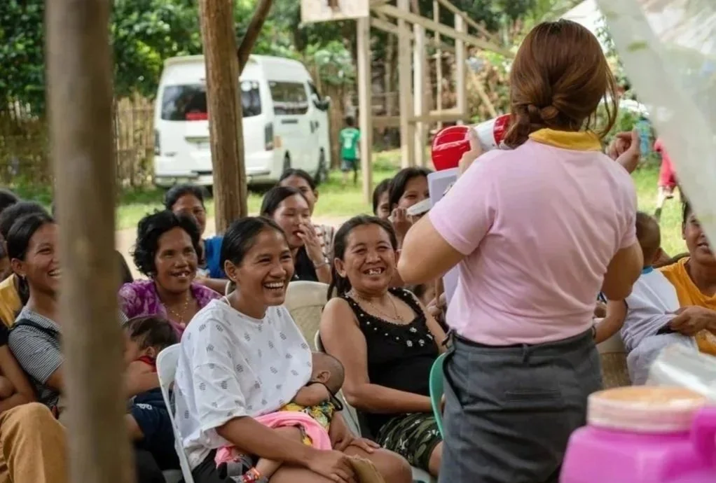 A woman in a pink shirt demonstrating something to a group of smiling women and children seated outdoors.