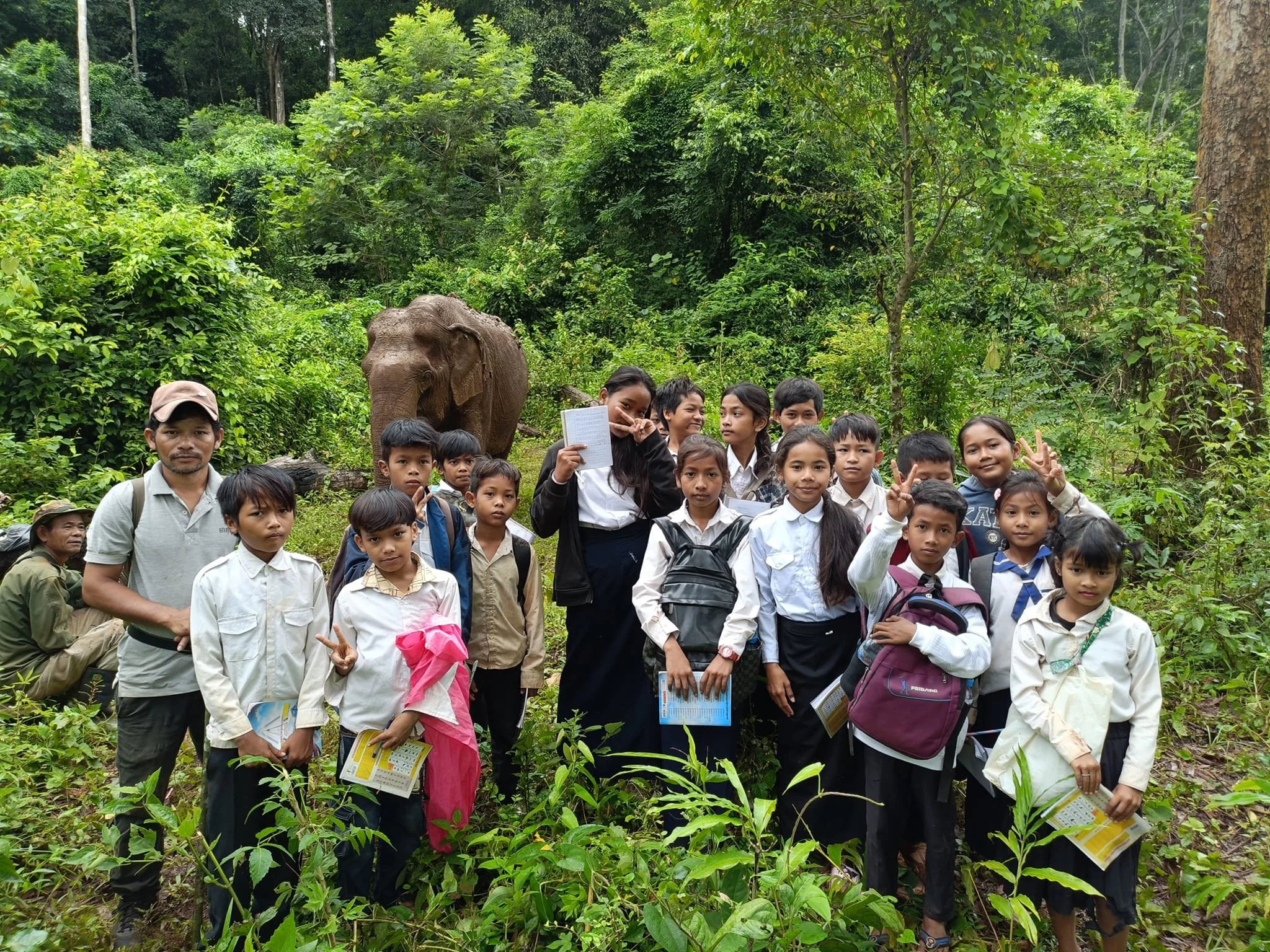 A group of children and adults posing with an elephant in a forest during an educational outing.