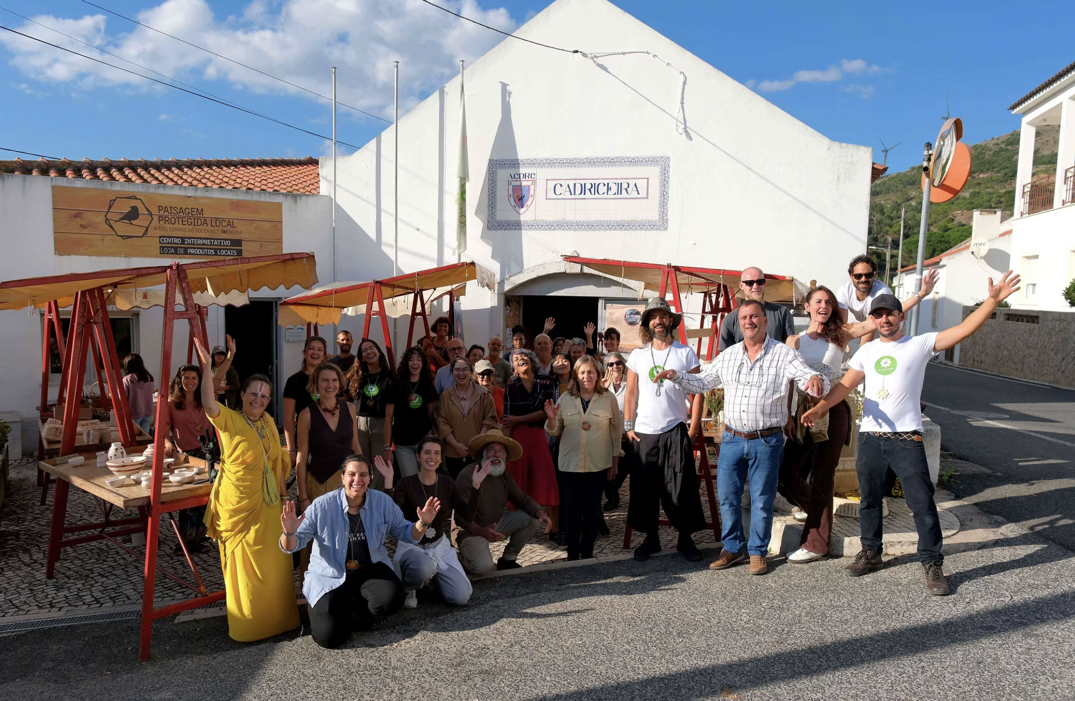 Group of people posing and smiling in front of a white building with sign that reads 'Cadriceira'. The scene is outdoors on a sunny day, with some people waving and others making gestures of celebration.