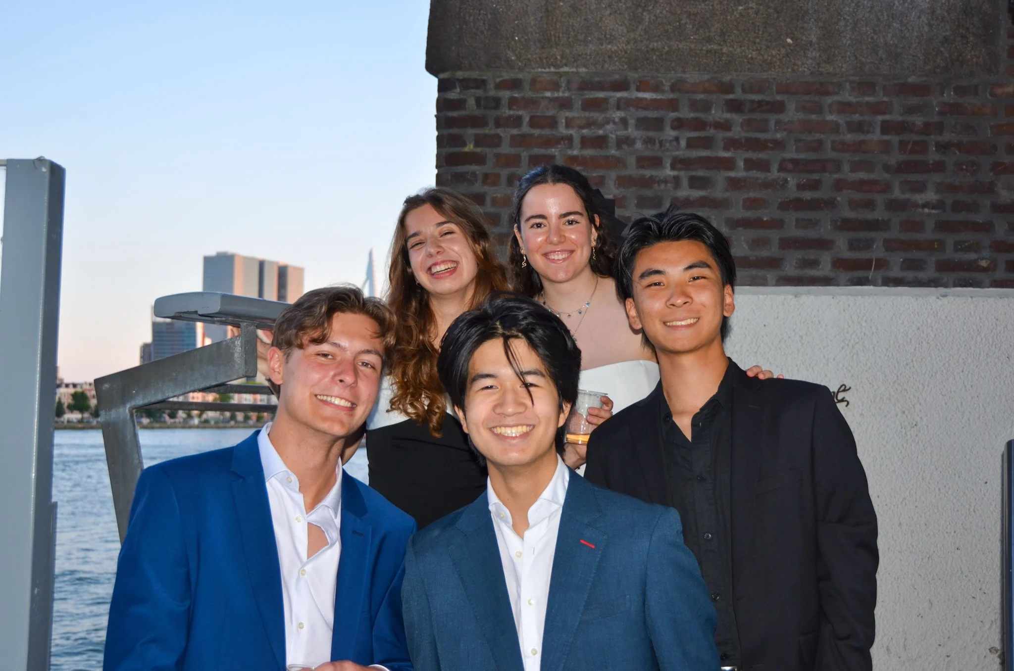 Five young adults in smart attire posing together outdoors against a brick wall.