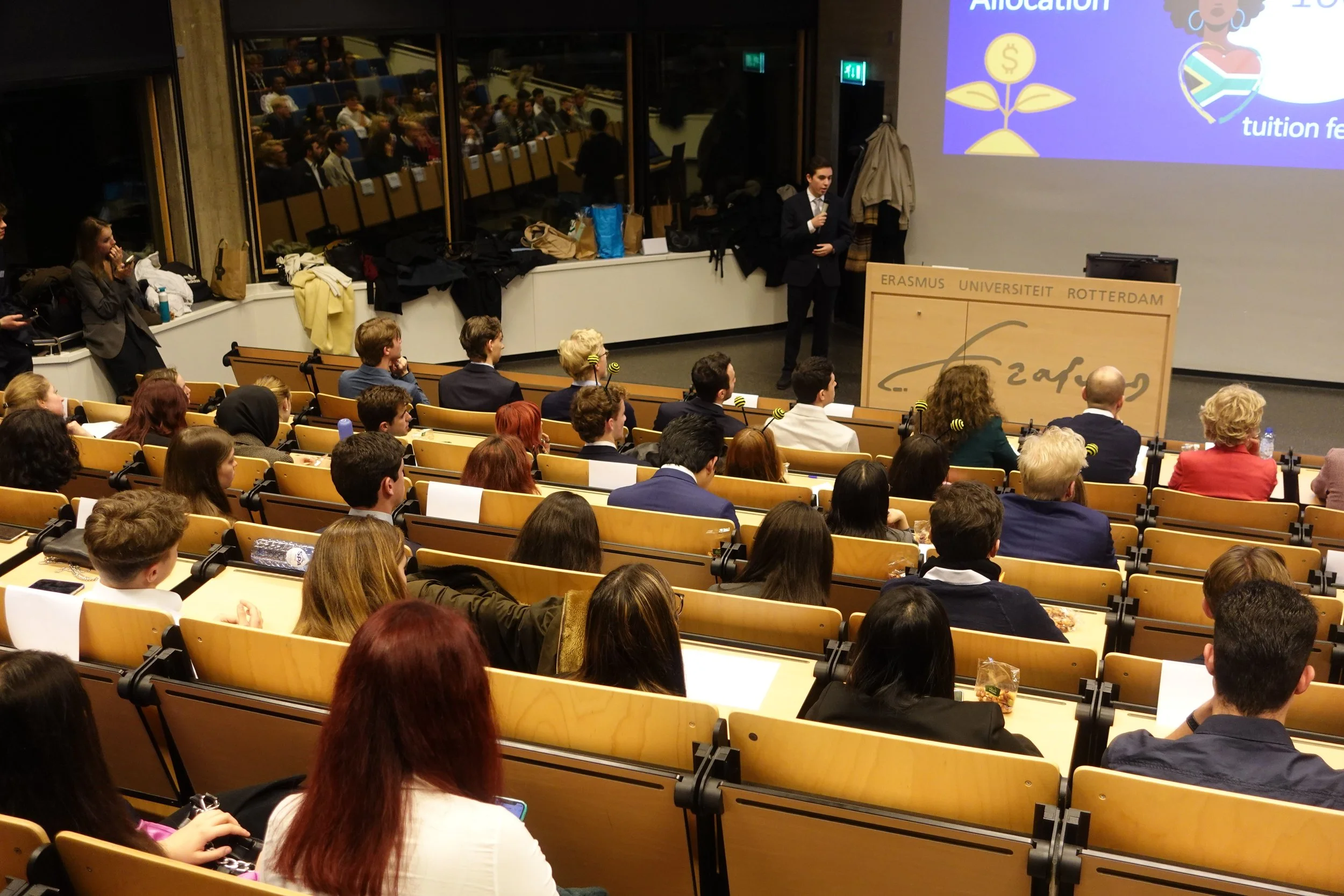 A lecture hall filled with students seated and listening to a presenter at the front. The presenter, dressed in a suit, stands near a podium with 'Erasmus Universiteit Rotterdam' written on it, and a large screen shows a presentation slide. Students in the audience are taking notes or watching attentively.