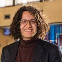 Portrait of a person with curly hair in business attire, standing outside with a blurred building.