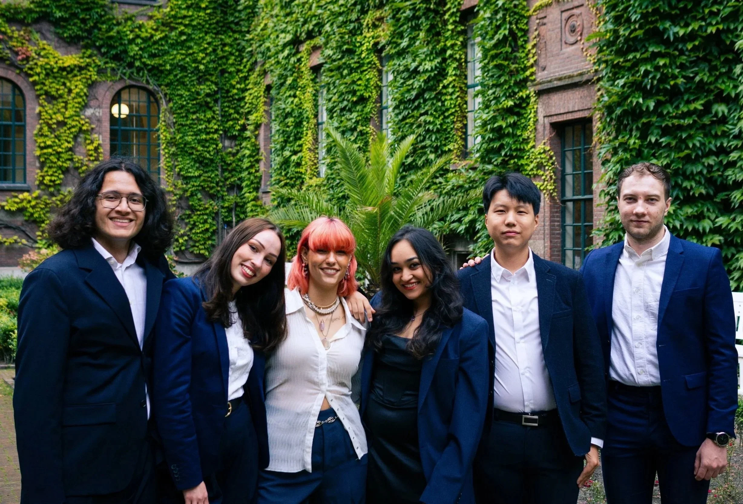 Six young adults in business attire posing together outdoors in front of an ivy-covered building.
