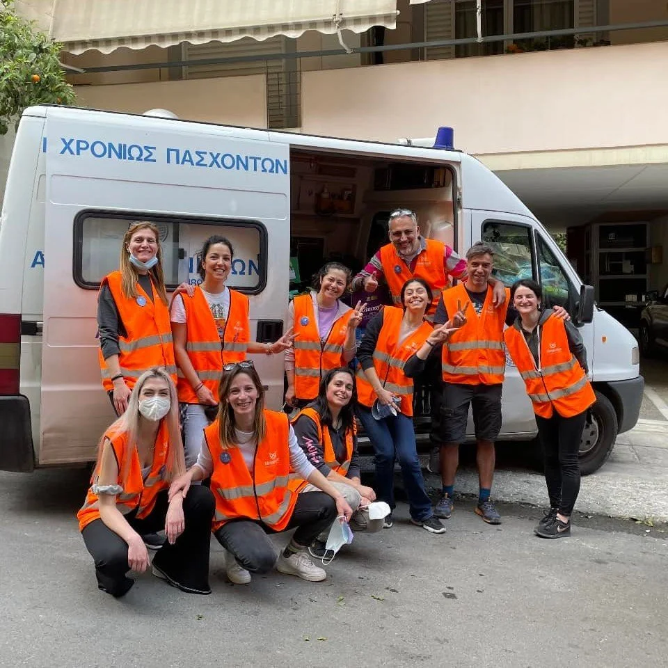 Group of people wearing orange safety vests posing for a photo in front of a white van with Greek writing on it, some standing and some kneeling, inside a parking lot.