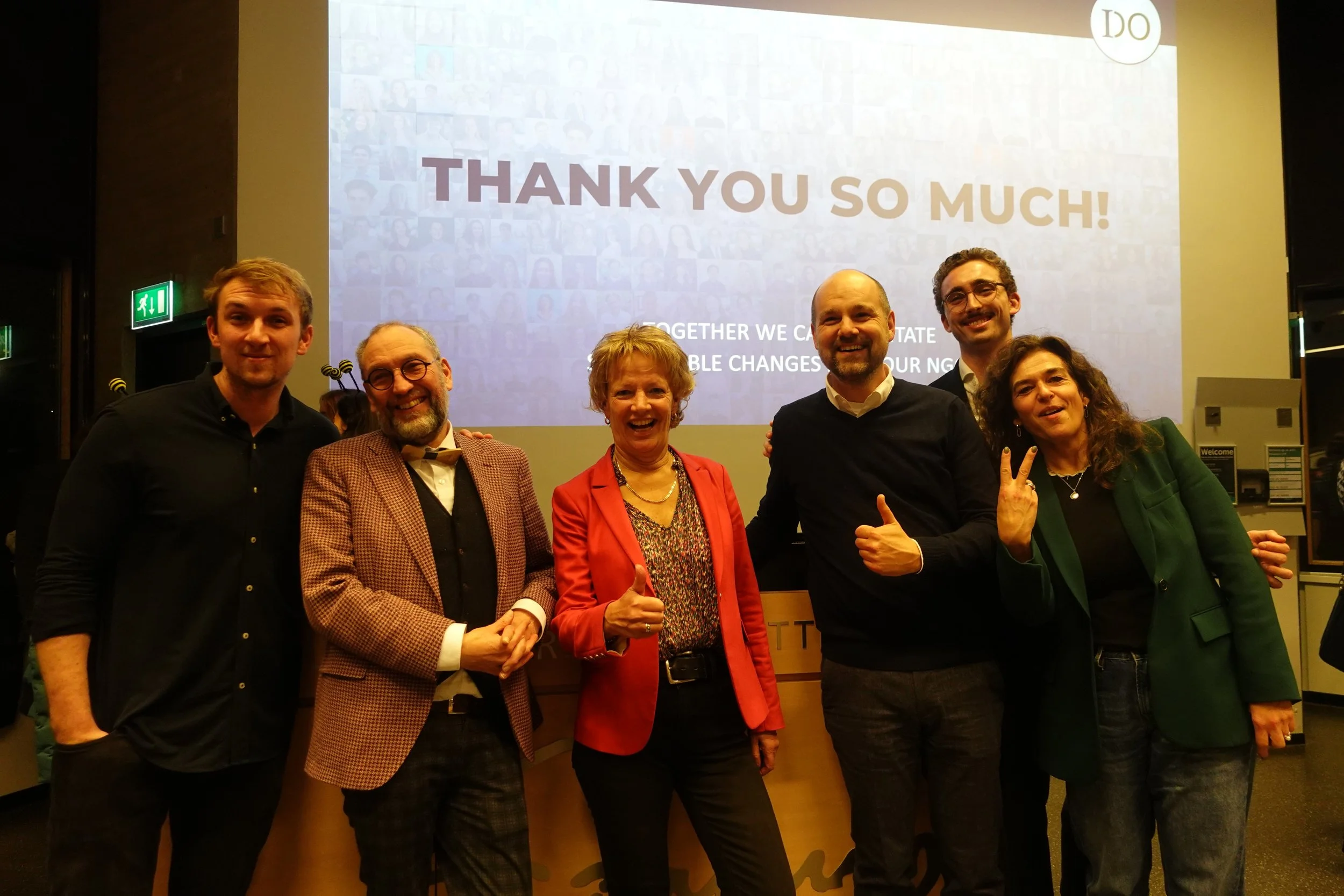 Group of six people smiling and posing in front of a large screen that says 'Thank You So Much!' with a collage of faces in the background