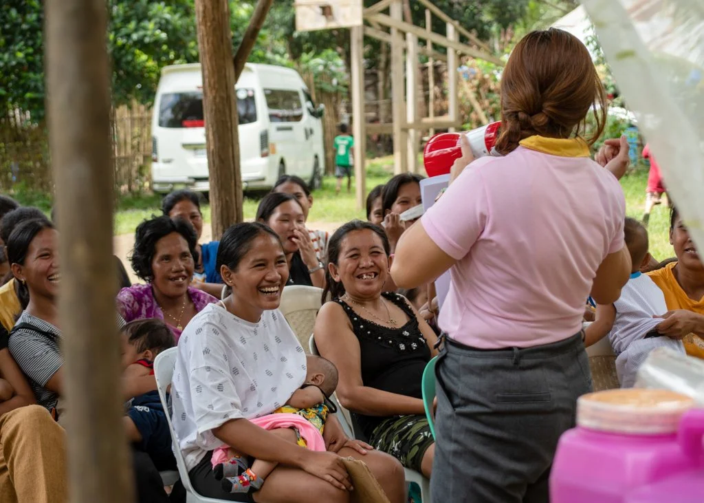 Community health worker with megaphone speaks to seated mothers, one breastfeeding her baby, during an outdoor gathering.