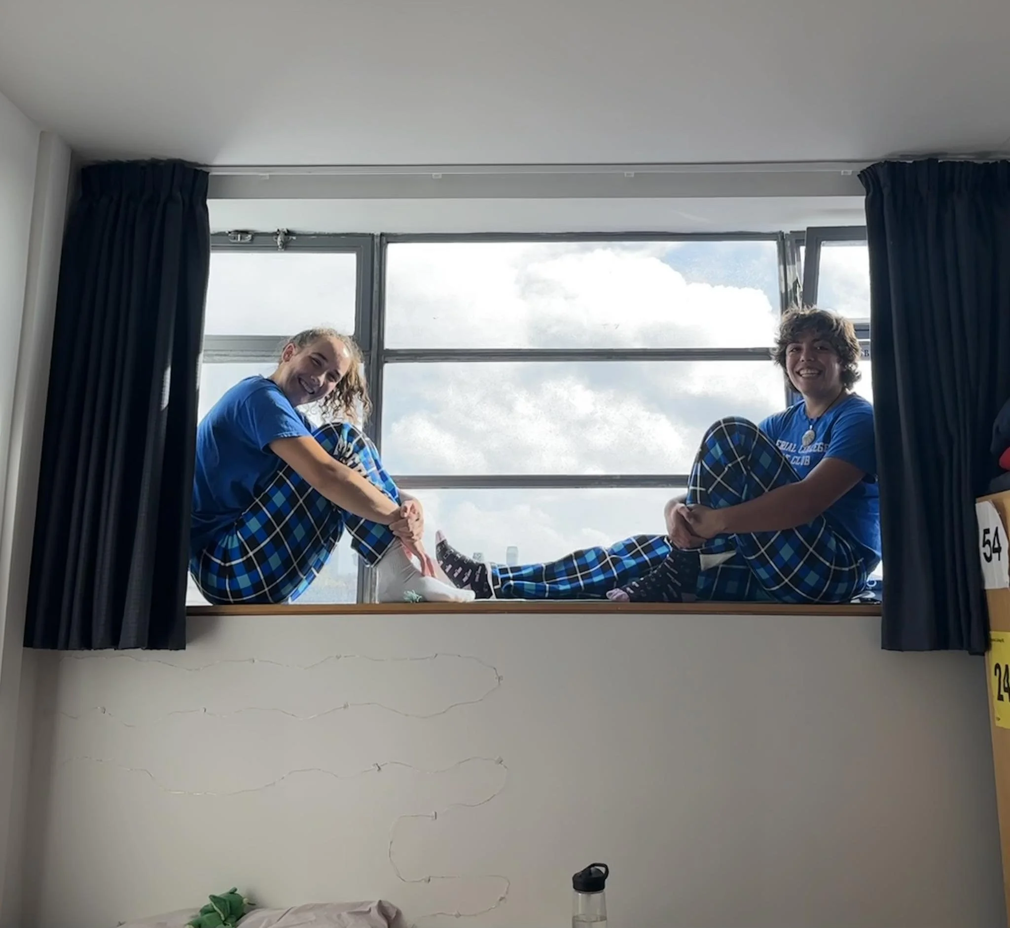 Two girls sitting on a window sill, wearing matching blue shirts and plaid pants, smiling at the camera with a cloudy sky in the background.