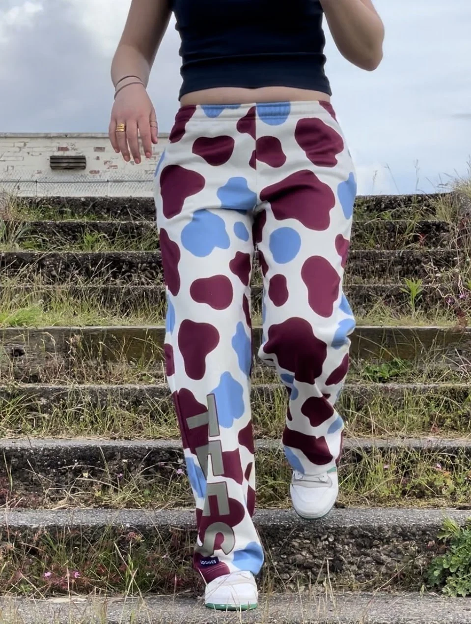 Person wearing cow-patterned pants and white sneakers, walking down outdoor stone steps with grass and plants around, overcast sky in background.