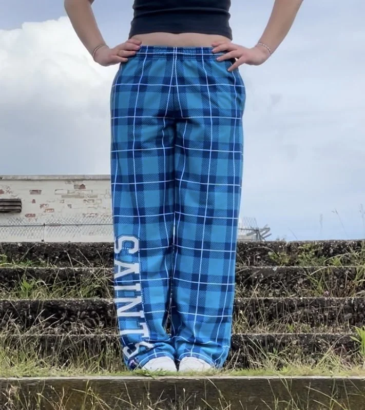 Person standing outdoors on steps, wearing blue plaid pajama pants with white text on the leg, a black top, and white shoes, with hands on hips and cloudy sky in the background.