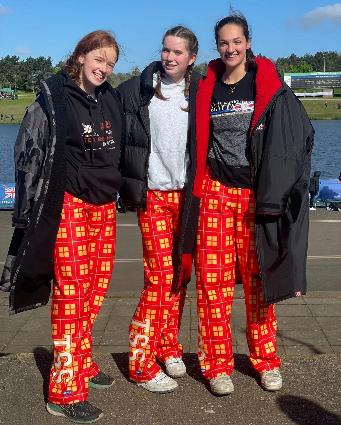 Three young women standing outdoors by a lake, smiling, wearing black jackets and red plaid pants with white lettering and designs, against a background of a sports field and clear blue sky.