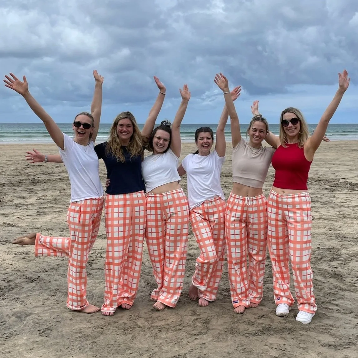 Six women wearing matching peach and white checkered pants, standing on a sandy beach with cloudy skies and the ocean in the background, celebrating with their arms raised and smiling.