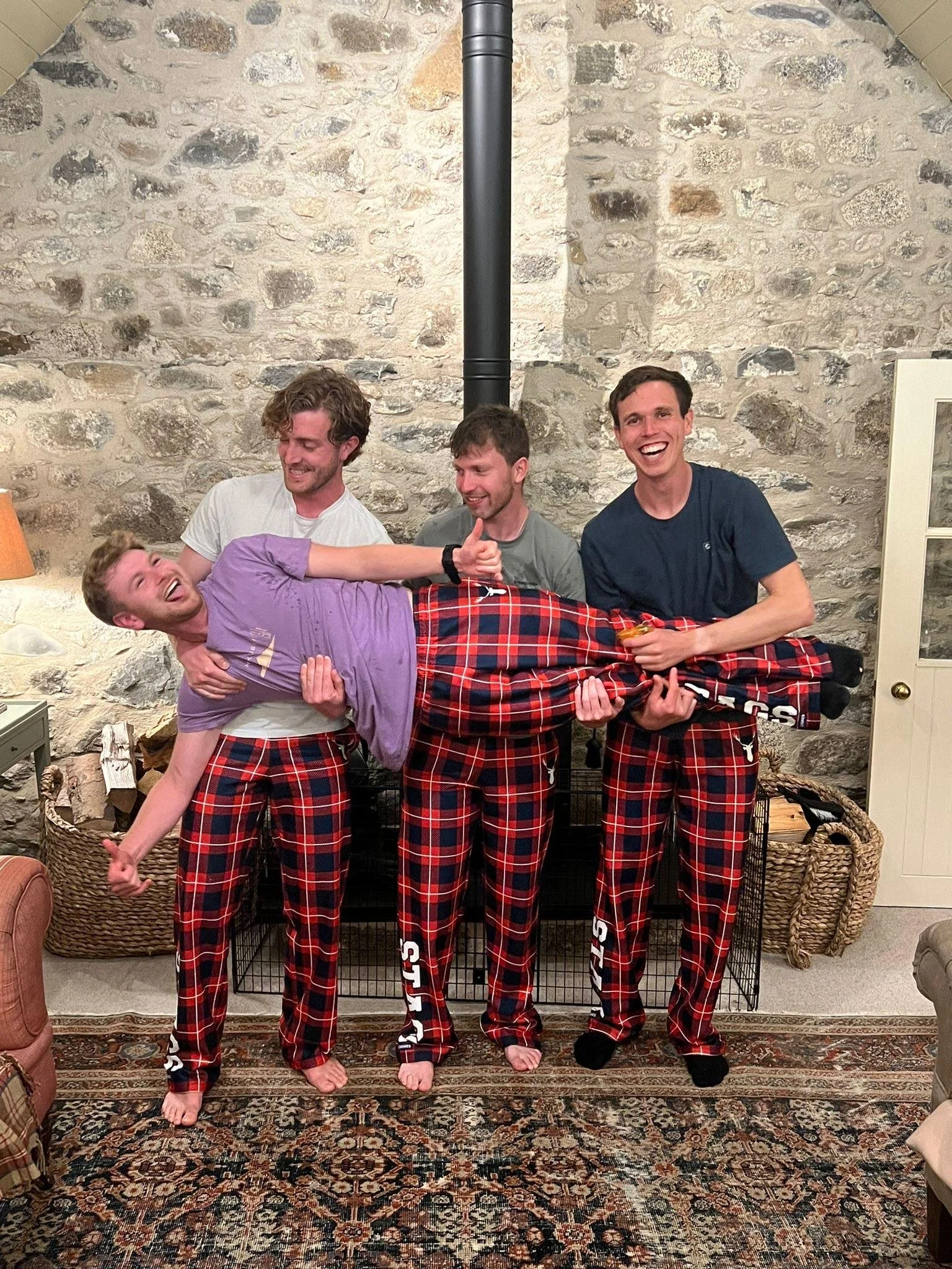 Four men are standing in front of a stone fireplace with a chimney pipe. They are wearing matching red and black plaid pajama pants. One man is being held horizontally by the other three men, all smiling and making playful gestures.