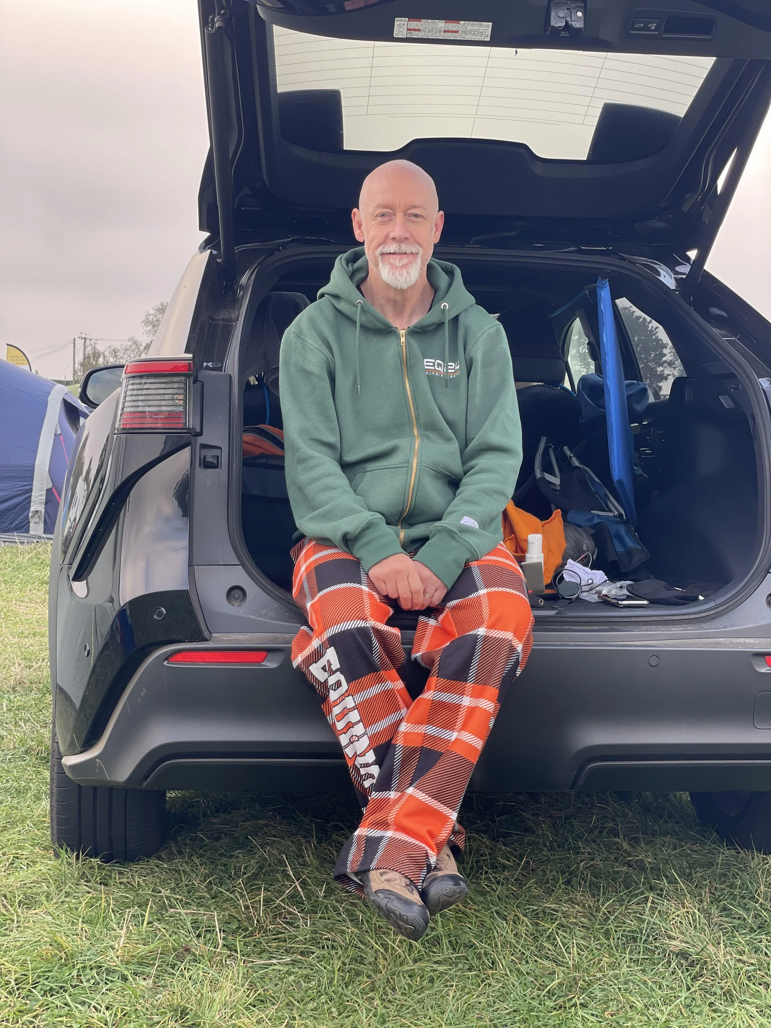 Man sitting on the back of an open SUV trunk outdoors, wearing a green hoodie and orange plaid pants.