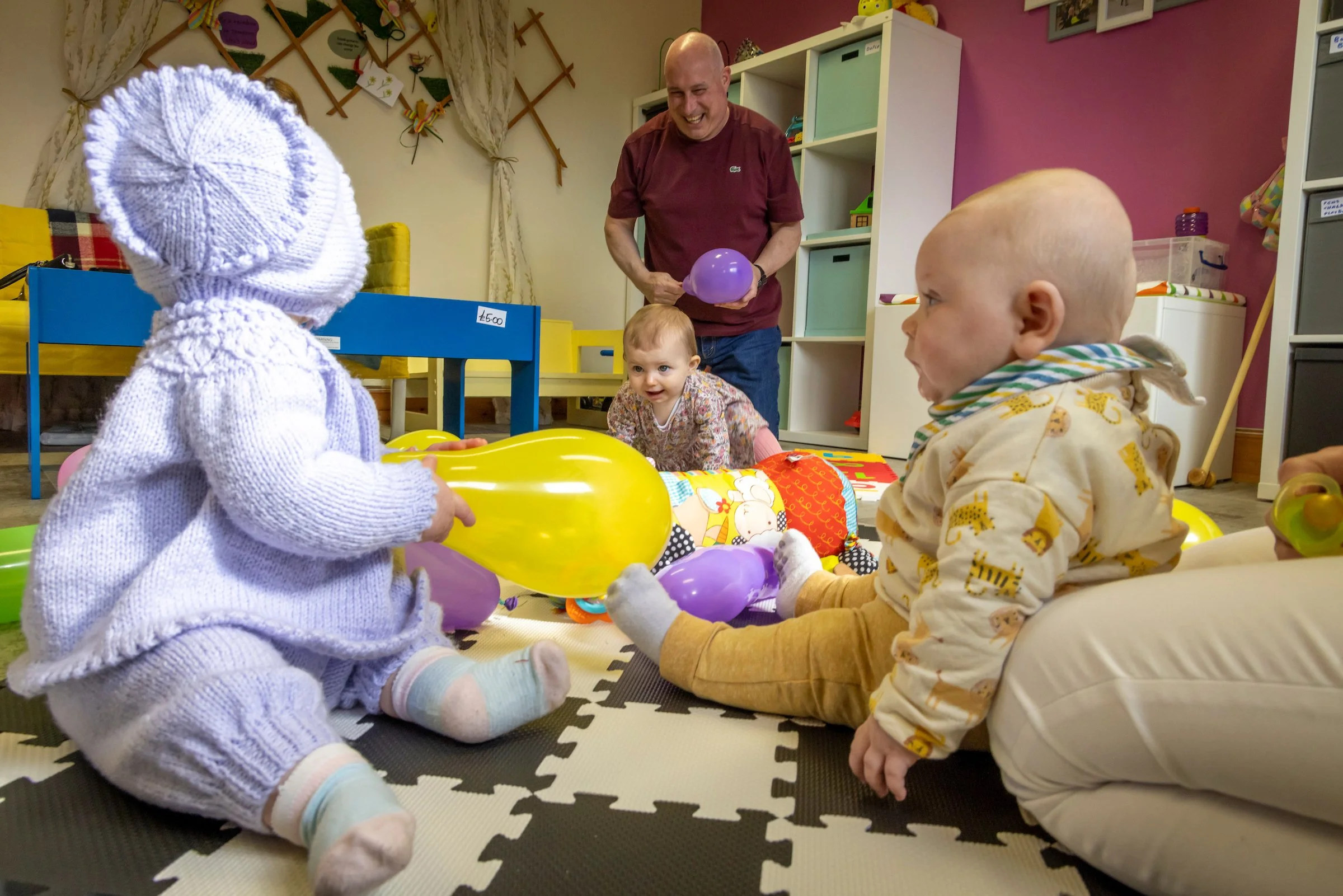 Children sitting on a foam mat playing with balloons, with a smiling adult man standing nearby in a colorful playroom.