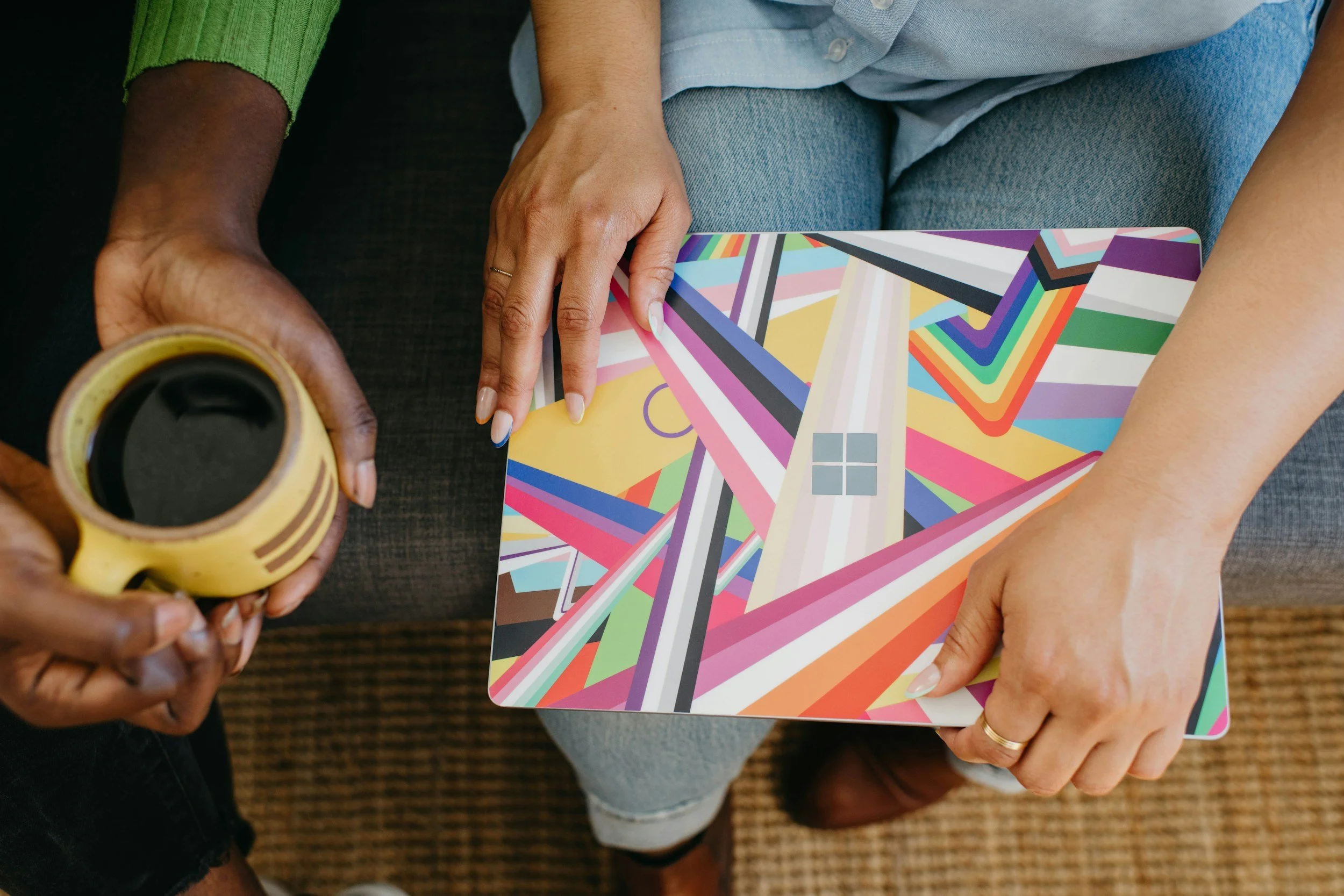 Two people sitting on a couch, one holding a yellow cup of coffee and the other holding a colorful, geometric-patterned tablet with a window illustration.