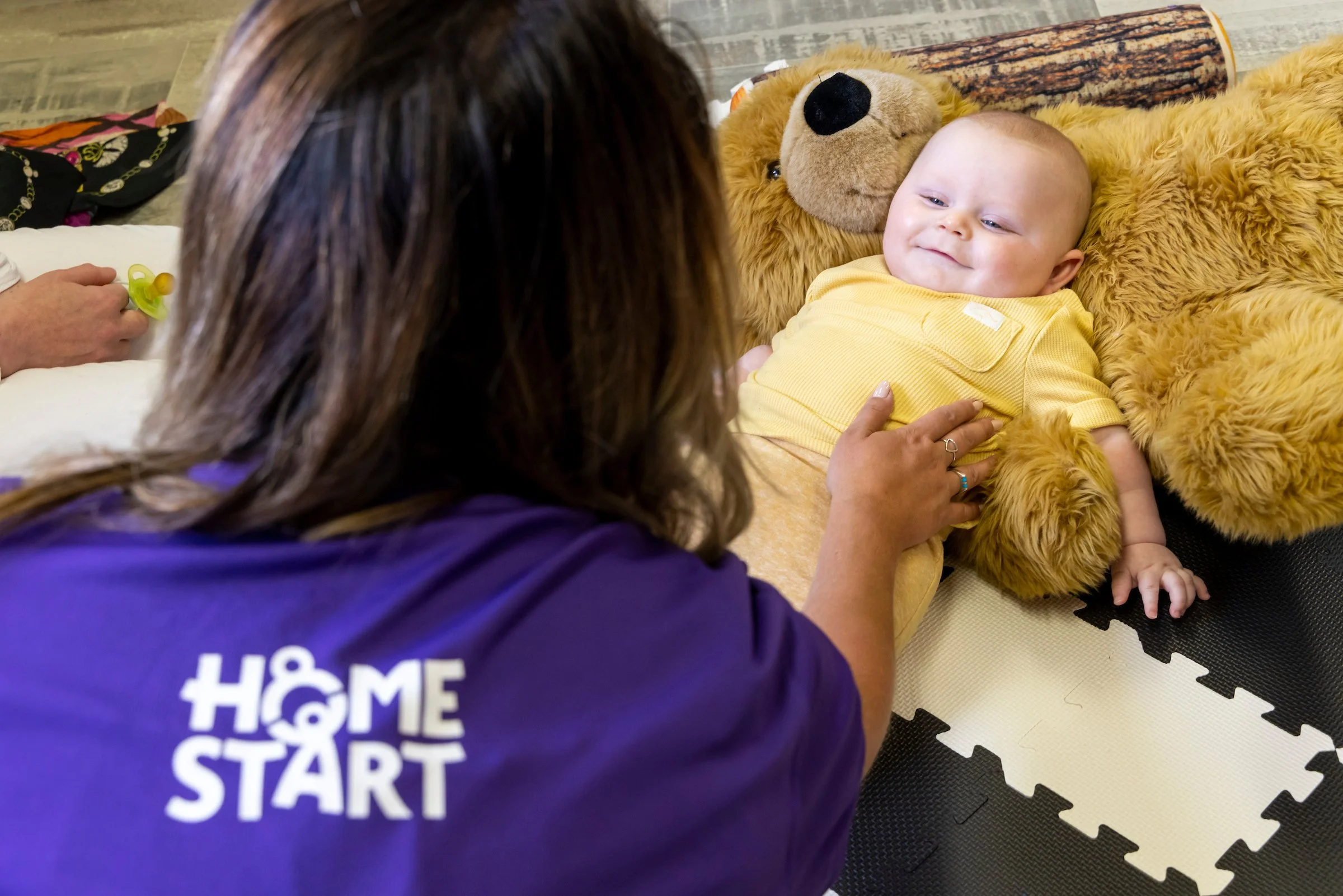 A woman wearing a purple shirt with 'Home Start' logo is gently touching a smiling baby lying on a teddy bear-themed blanket. The baby is dressed in a yellow outfit and is lying on a gray foam mat.