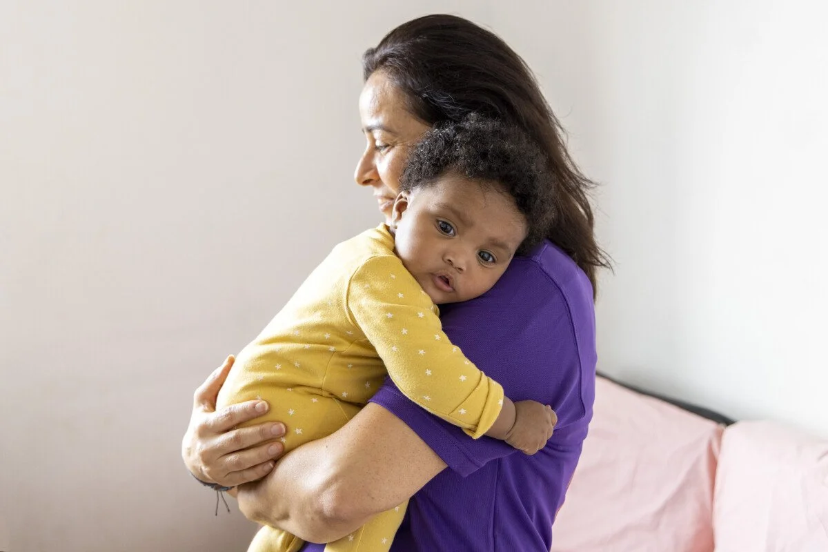 A woman hugging an African American child in a yellow outfit with white stars, against a plain light-colored wall.