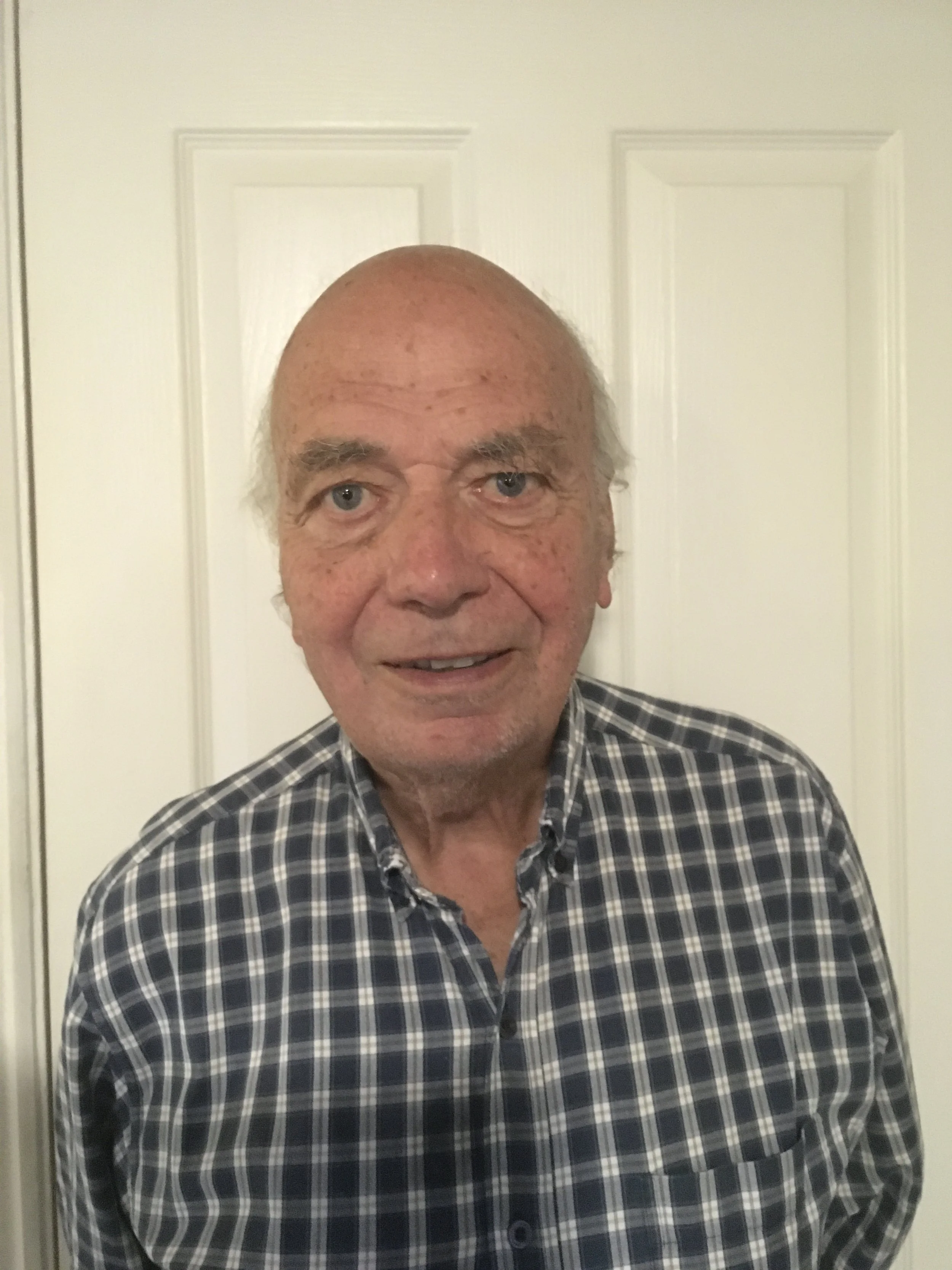 Home-Start trustee, A smiling elderly man with thinning hair and a bald spot, wearing a plaid shirt, standing in front of a white door.