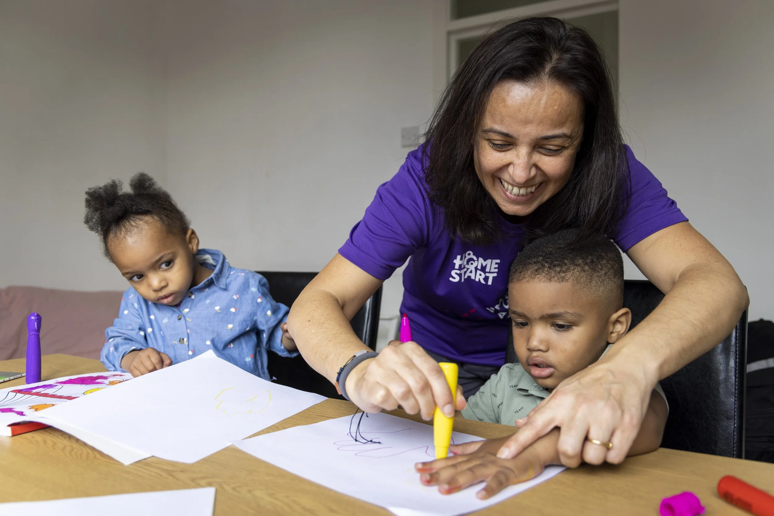 A woman with shoulder-length dark hair, smiling, helping a young boy draw on paper with a yellow marker at a table. A young girl with curly hair, wearing a blue shirt, looks on with a curious expression in the background.