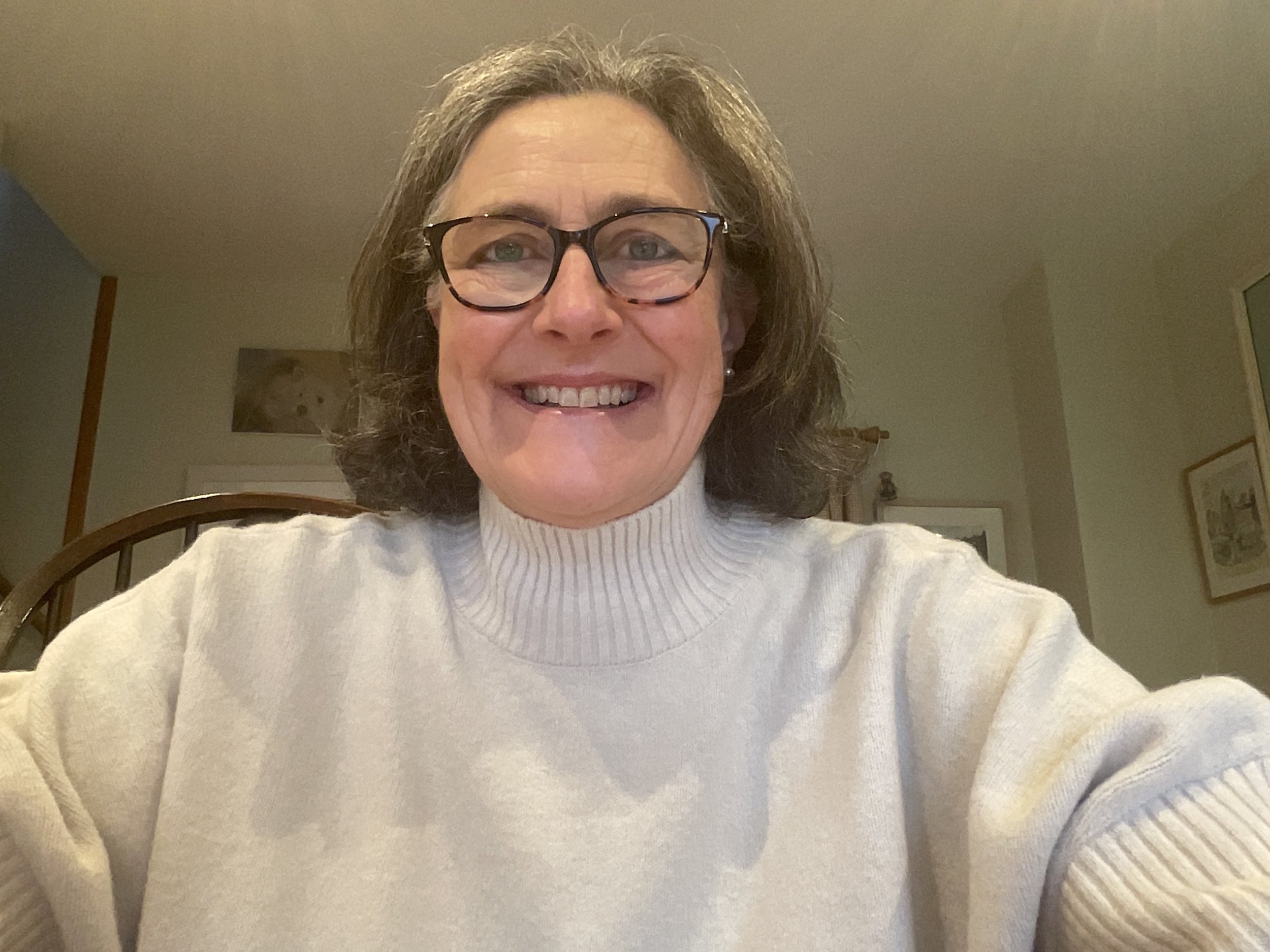 Home-Start trustee, A woman with shoulder-length hair, glasses, and pearl earrings, smiling and wearing a cream-colored turtleneck sweater, sitting indoors with framed pictures on the wall behind her.