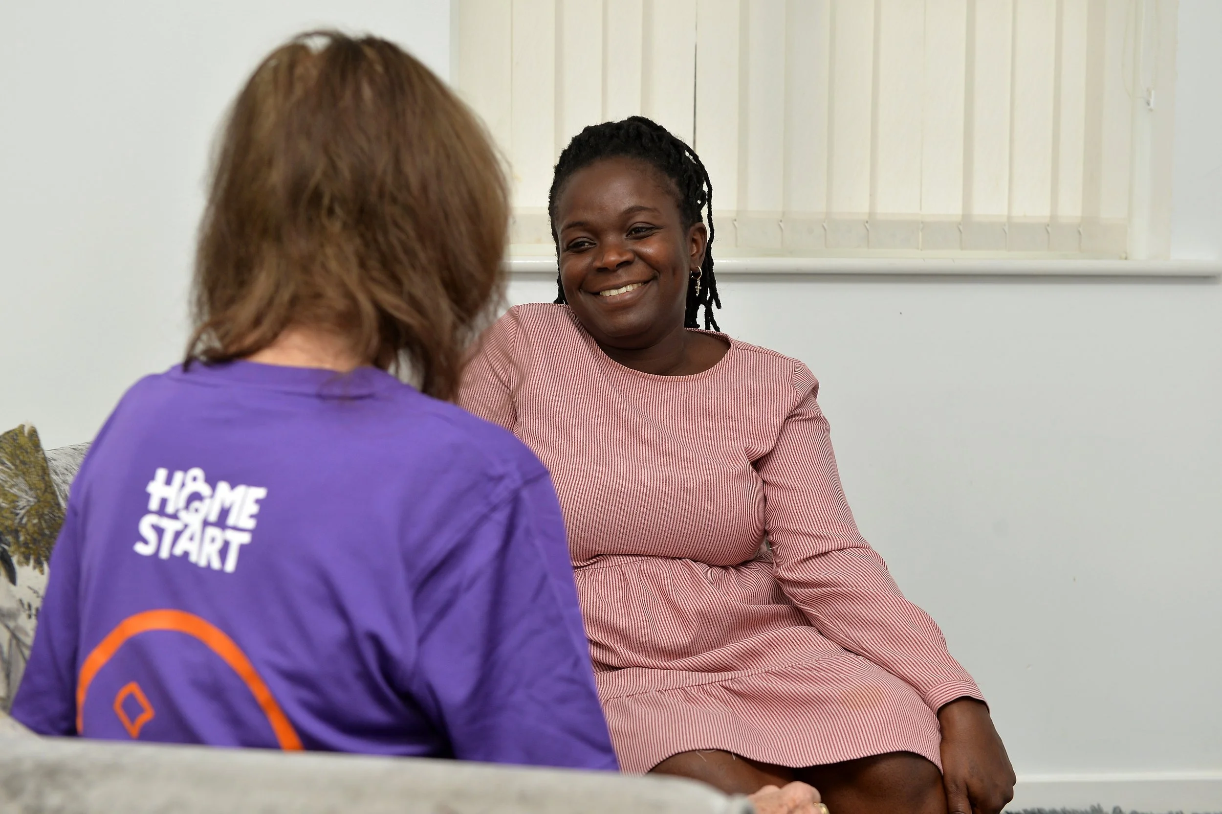Two women sitting on a couch, engaging in a friendly conversation indoors. The woman on the right is smiling and wearing a pink and white striped dress. The woman on the left has her back to the camera, with brown hair and a purple shirt with white text that says 'HOME START' on the back.
