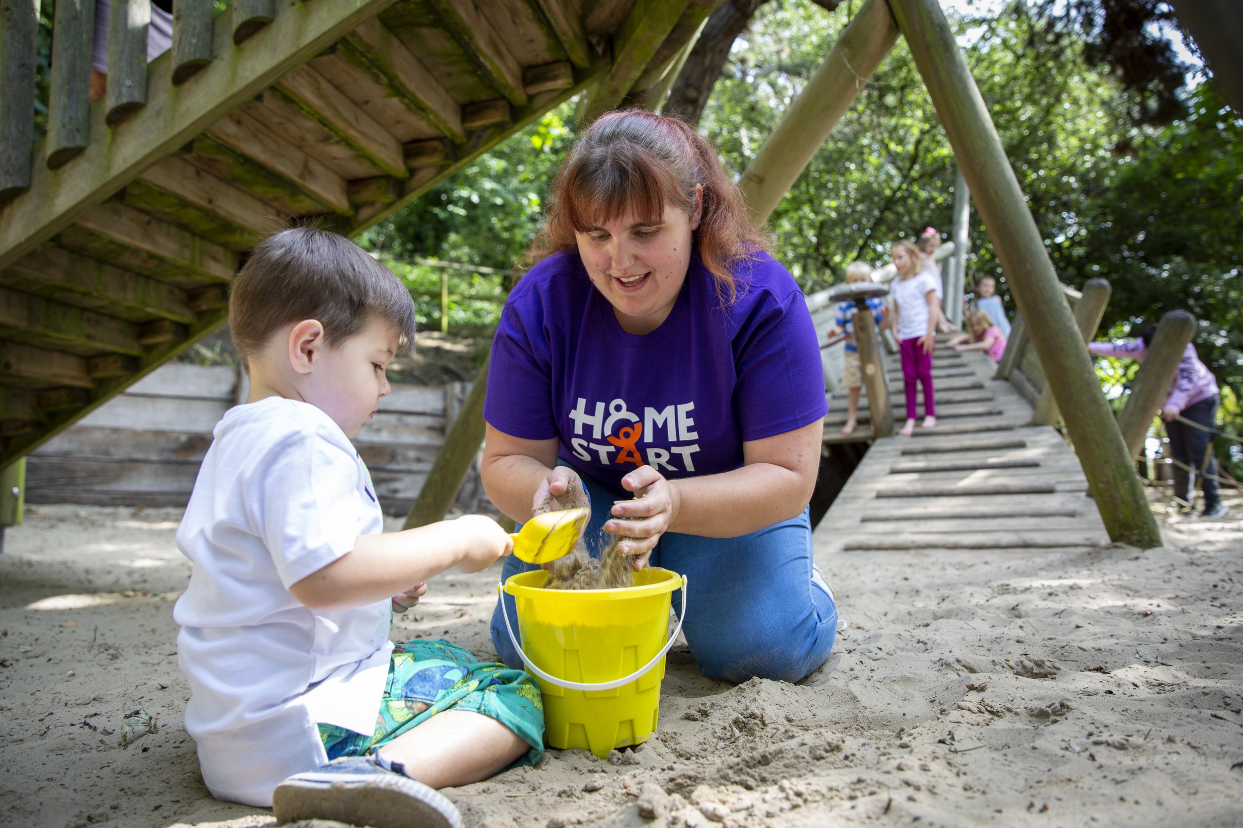 A home-start volunteer and a young boy playing in the sand at a playground with children in the background climbing a wooden ramp.