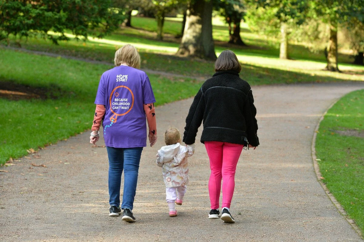 Three people, two women and a young girl, walking in a park on a paved pathway surrounded by green grass and trees.