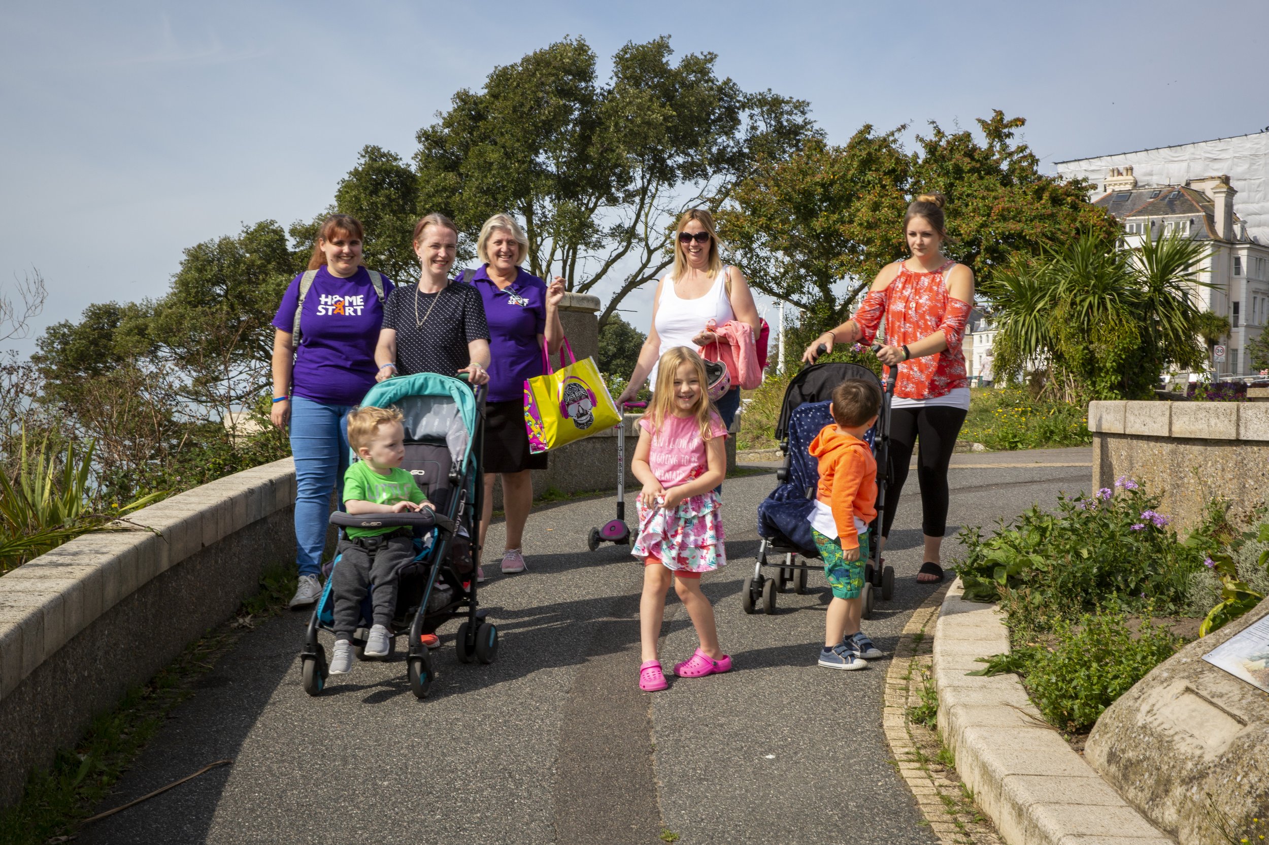 Group of women and children walking outdoors on a sunny day, some with strollers, on a paved path with greenery and trees in the background.