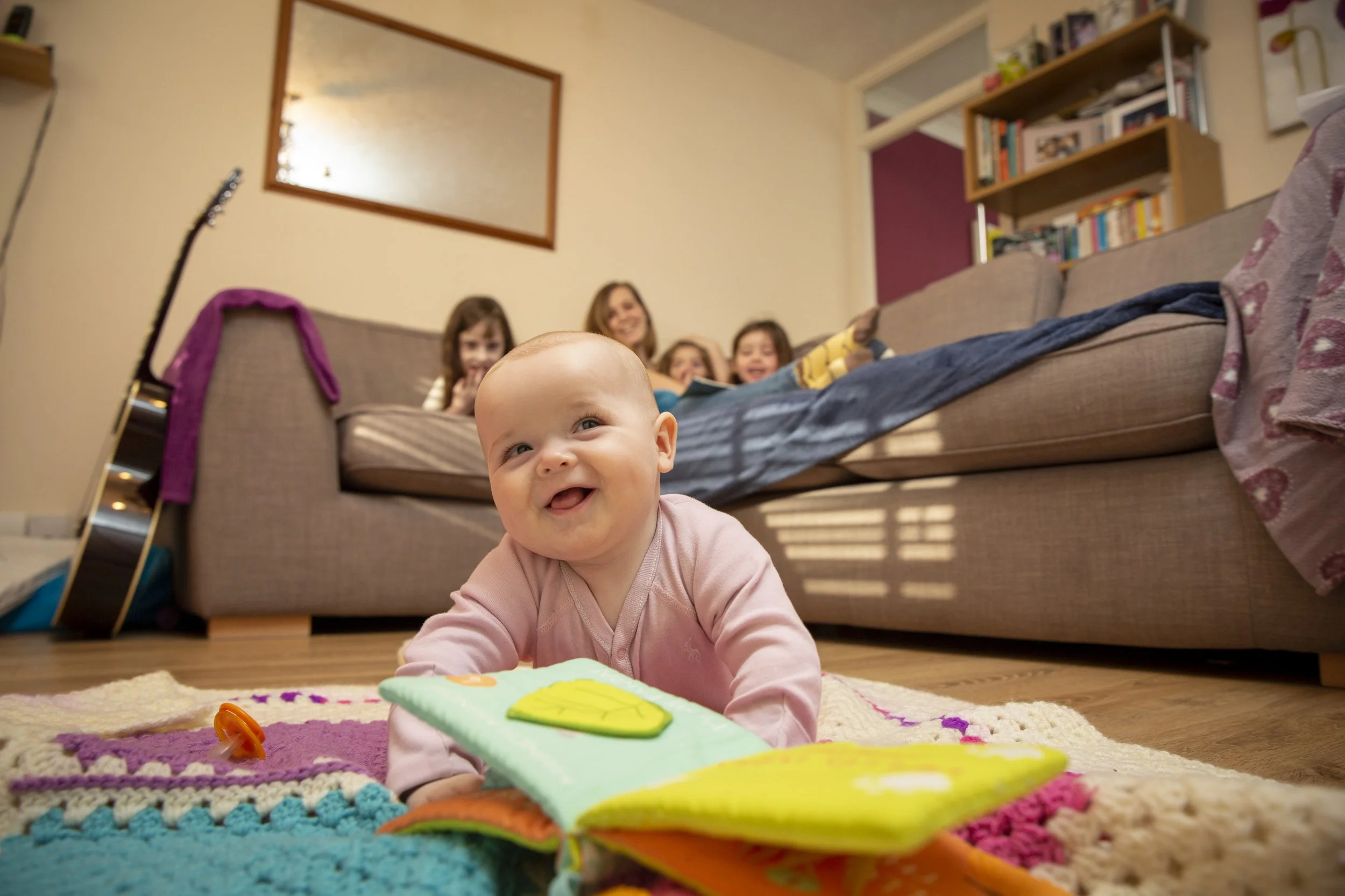 A baby lying on a colorful rug, smiling at the camera with an open book or fabric in front, with four children and an adult sitting on a couch in the background, inside a cozy living room.