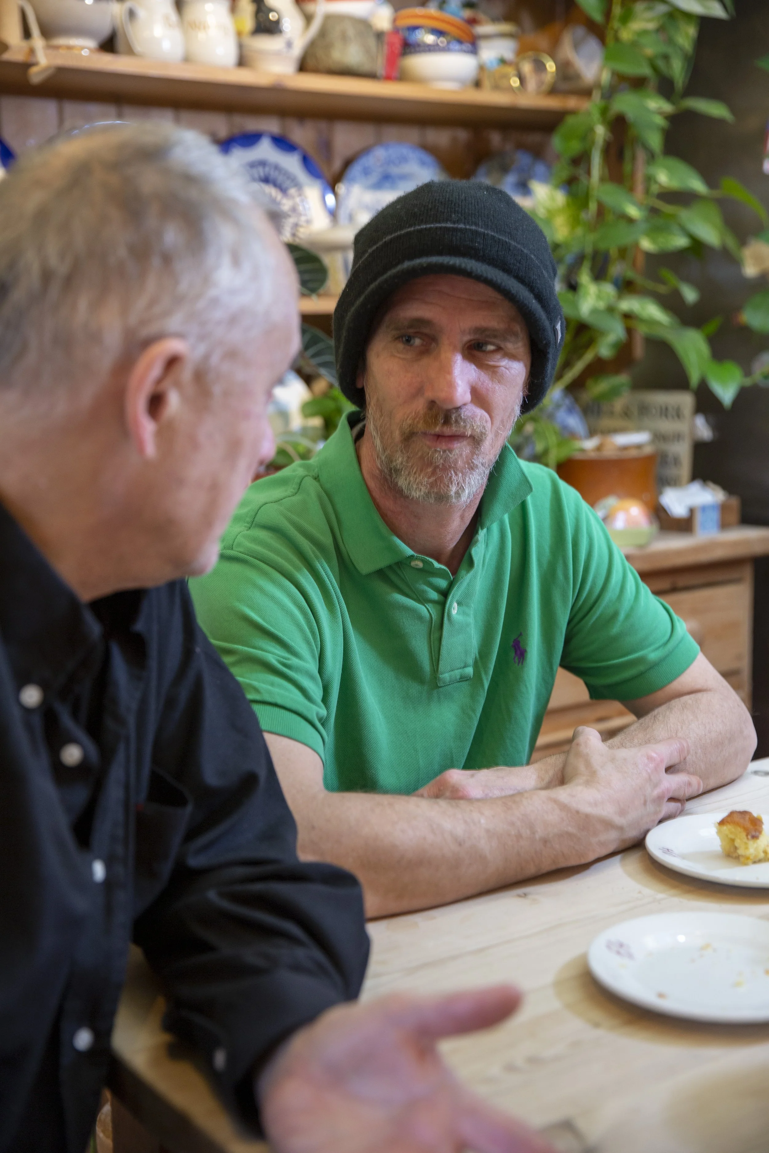 Two men sitting at a wooden table having a conversation; one wearing a green polo shirt and black beanie, the other with gray hair in a black shirt, with a plate of cake in front of them. Shelves with dishes and a green plant are in the background.