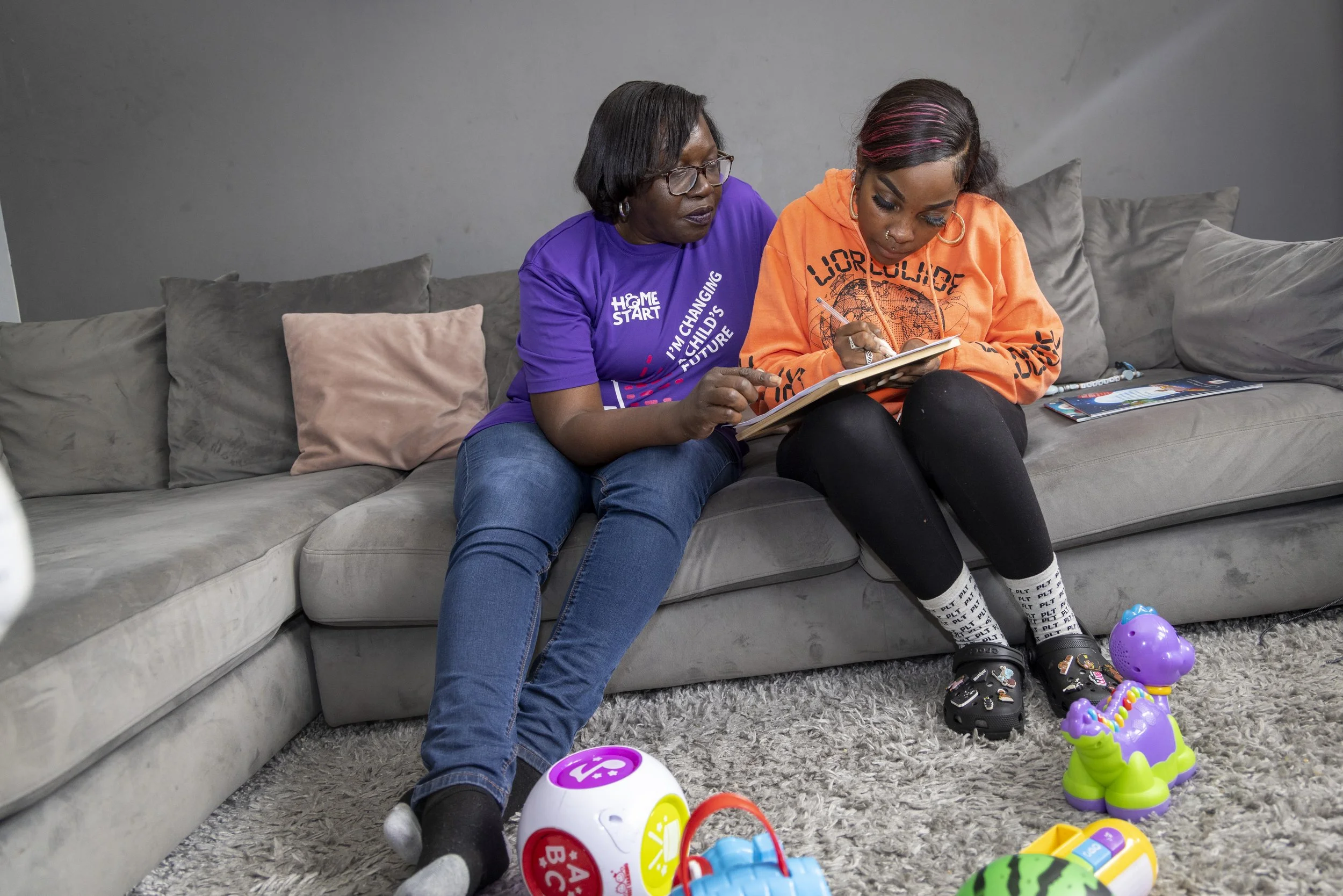 Two women sitting on a gray couch with toys on a gray carpet. The older woman is wearing a purple shirt and glasses, pointing at a notebook held by the younger woman who is dressed in an orange hoodie and black leggings.