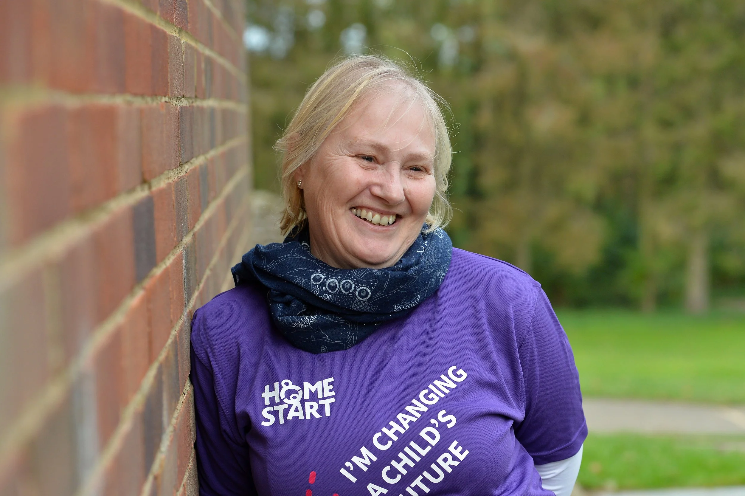Smiling woman leaning against a brick wall, wearing a purple shirt with the words 'HOME & START' and 'I'M CHANGING A CHILD'S FUTURE' printed on it, outdoors with trees in the background.