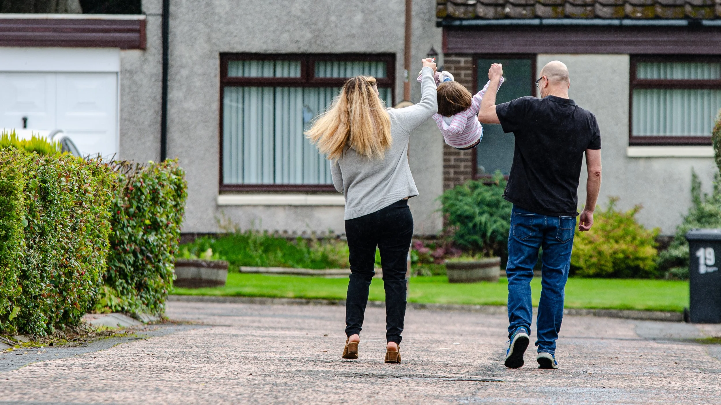 Family of three, two adults and a child, walking on a paved sidewalk outside their house. The woman and man are swinging the child by holding their hands.