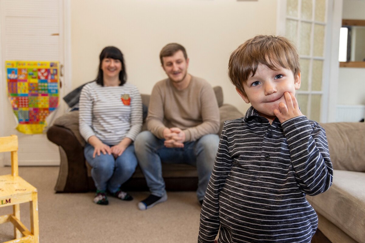 A young boy in a black and gray striped shirt with light brown hair standing in front of a beige couch, with a woman and a man sitting on the couch smiling in the background.