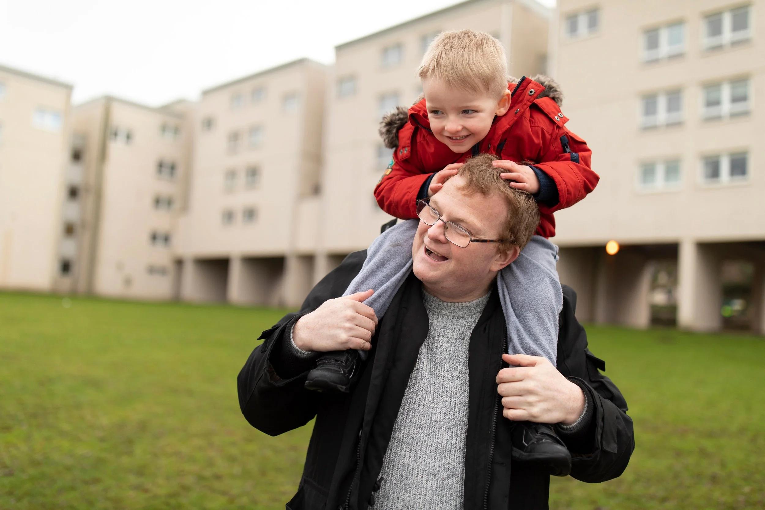 A man with glasses carrying a young boy on his shoulders in a grassy area in front of a beige apartment building. The boy is smiling and wearing a red jacket.