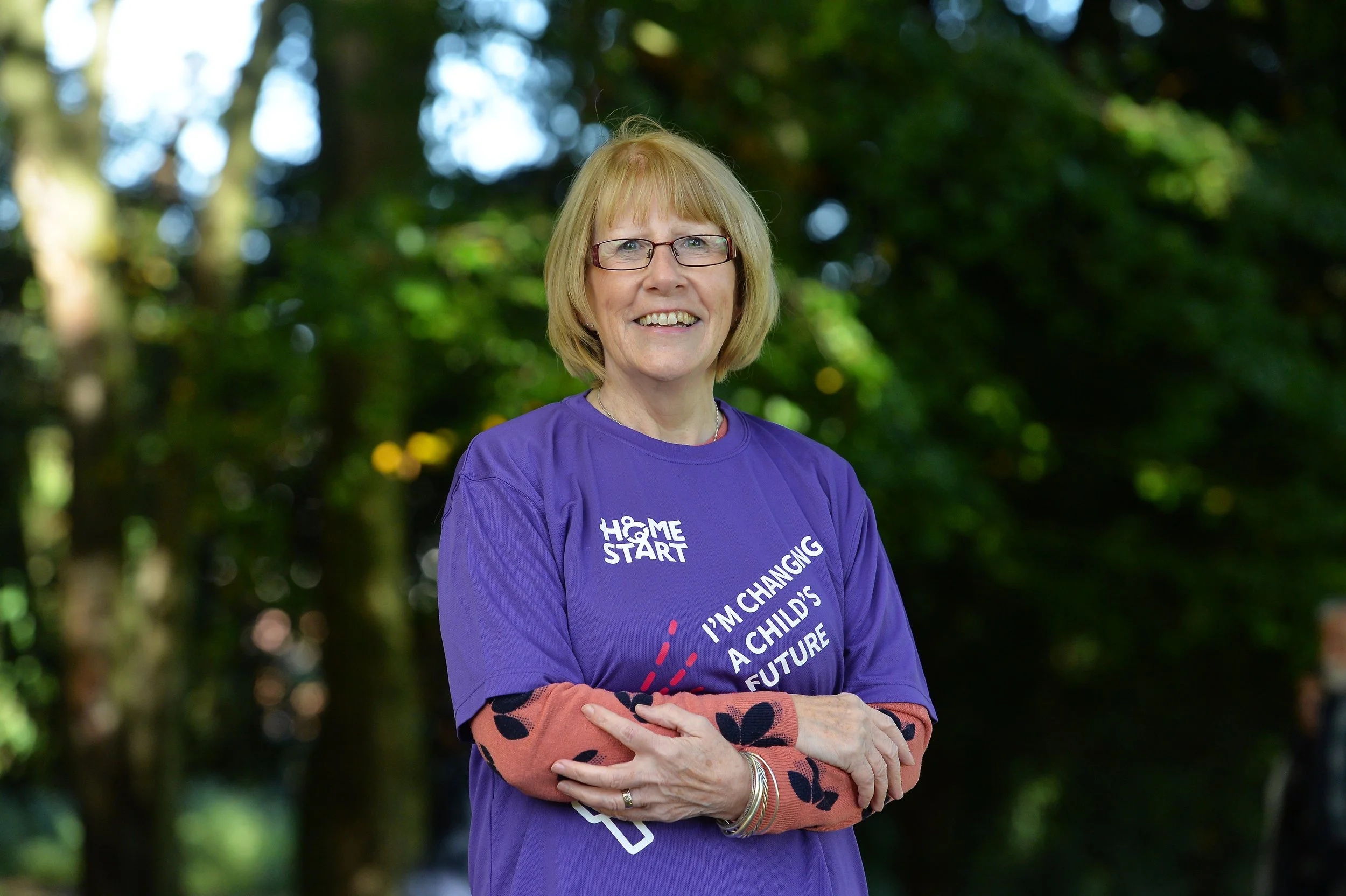 A smiling elderly woman with glasses and short blonde hair stands outdoors in front of green foliage, wearing a purple t-shirt with white text and an orange long-sleeve shirt underneath.