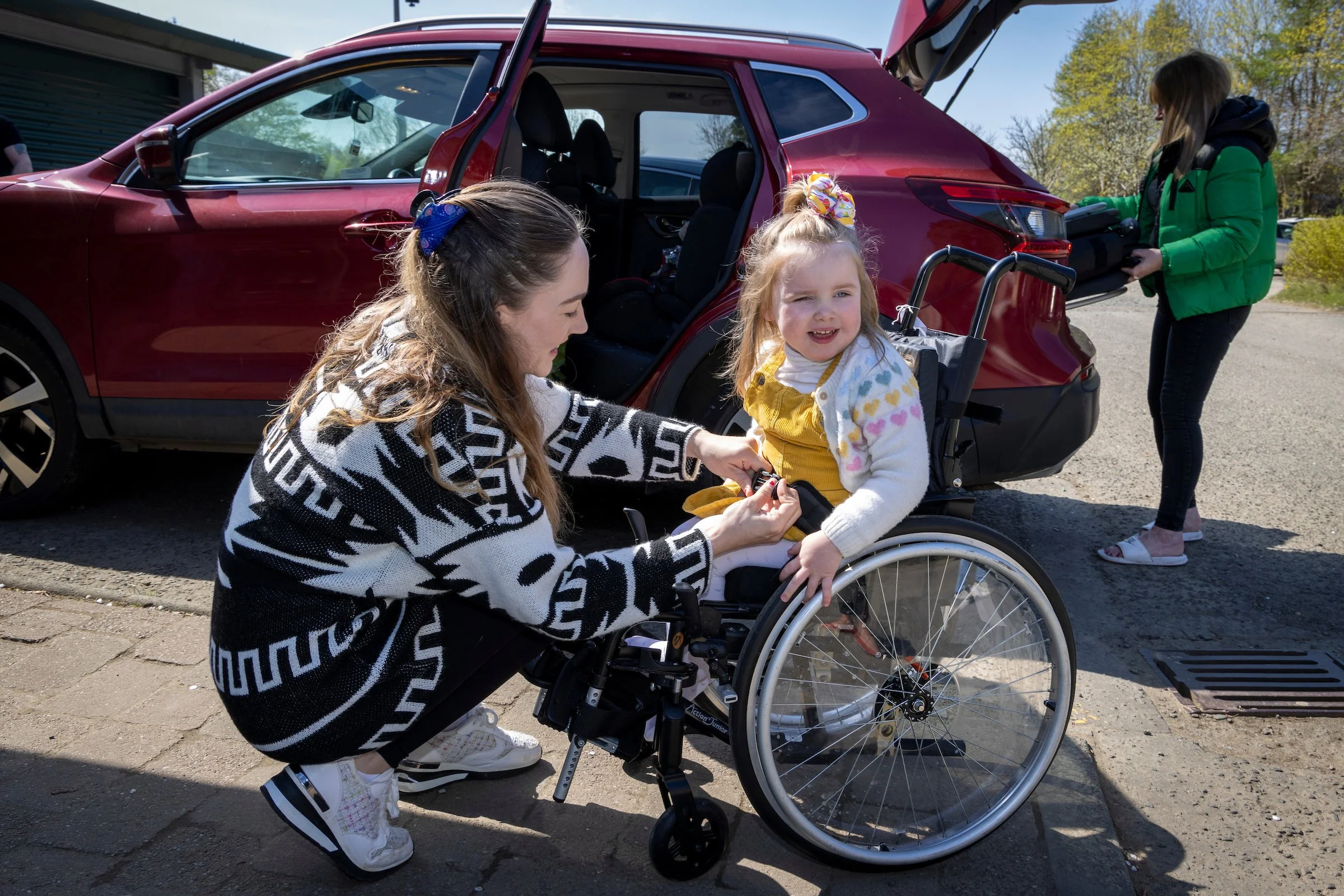 A young girl in a wheelchair smiling, with a woman adjusting her clothing, beside a red SUV with an open trunk. In the background, another woman is retrieving luggage from the car on a sunny day with trees lining the street.
