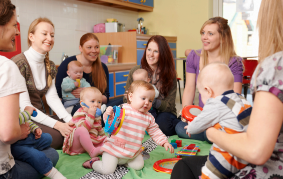 Group of young women and toddlers engaging in a play session on a green mat in a colorful classroom.