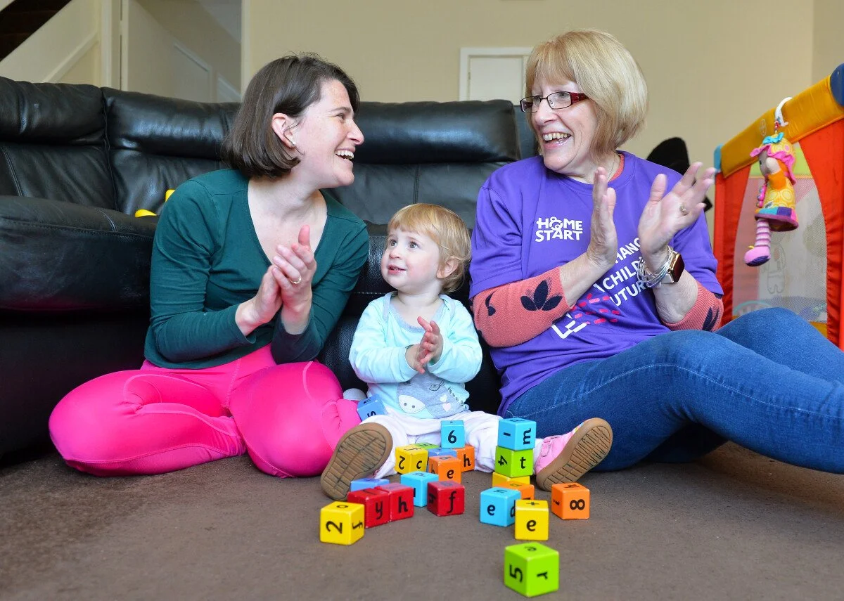 Three people, an adult woman, a young girl, and an older woman, sit on the carpeted floor playing with colorful alphabet blocks. They are smiling and clapping, enjoying a happy moment together in a cozy room.