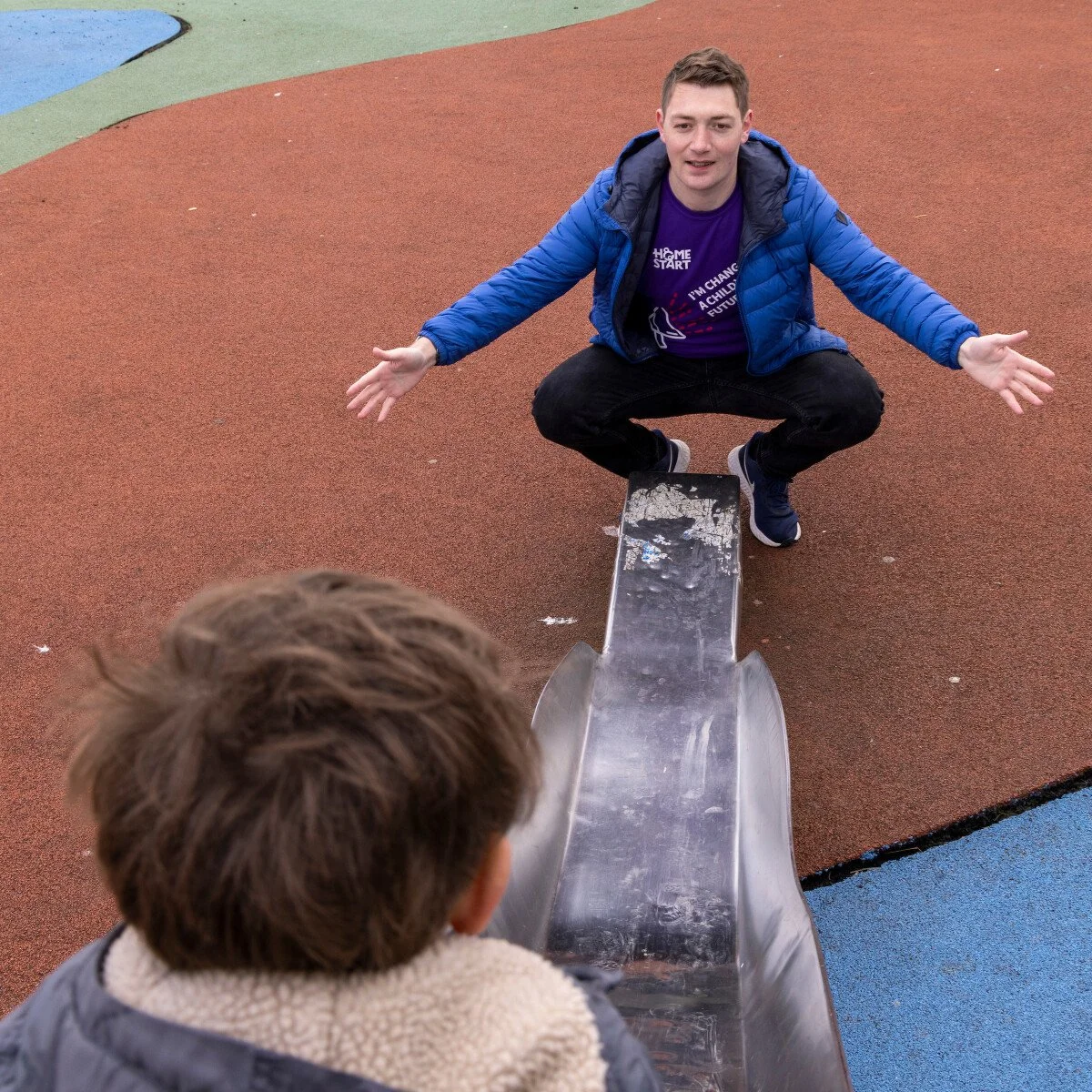 A man squatting on a colorful playground surface with arms outstretched, facing a child at the top of a metal slide.