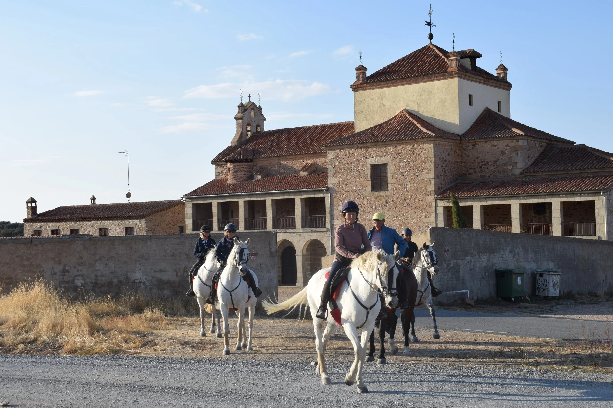 People riding horses in front of a historic stone building with a tiled roof, under a clear blue sky.