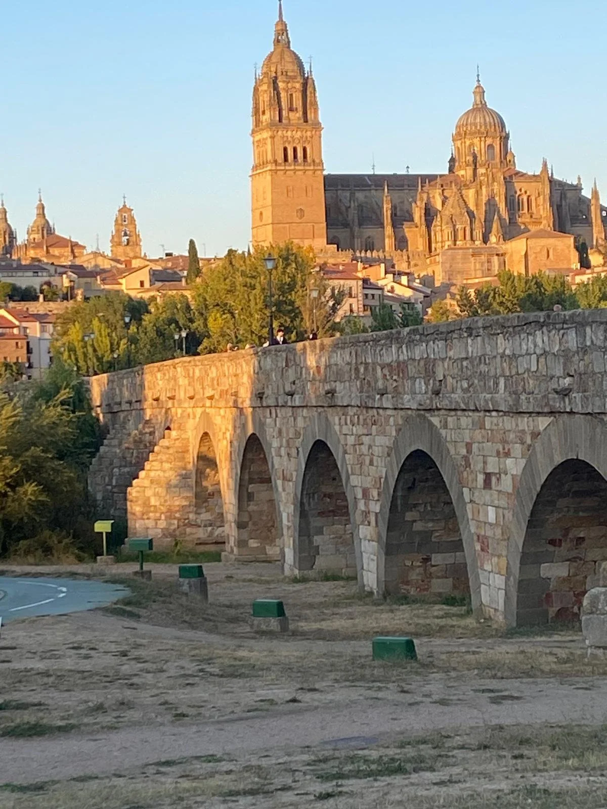 Sunset view of an ancient stone bridge with arches, overlooking a city with historic cathedrals and buildings.
