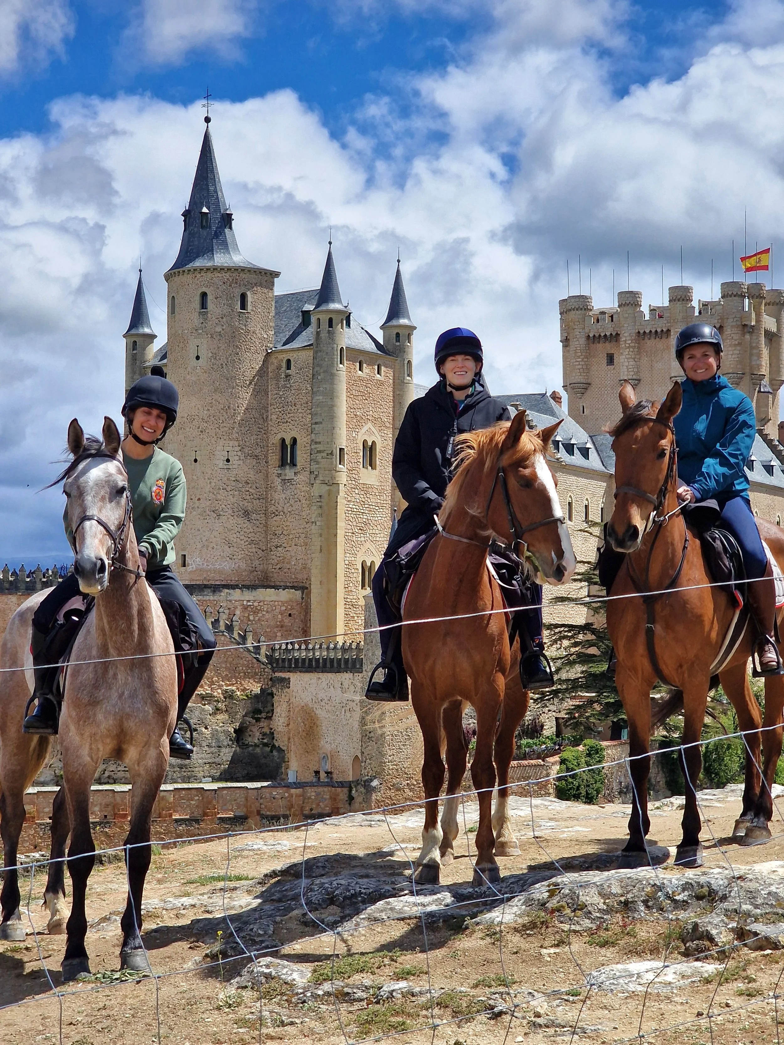 Three women riding horses in front of a large medieval castle on a partly cloudy day.