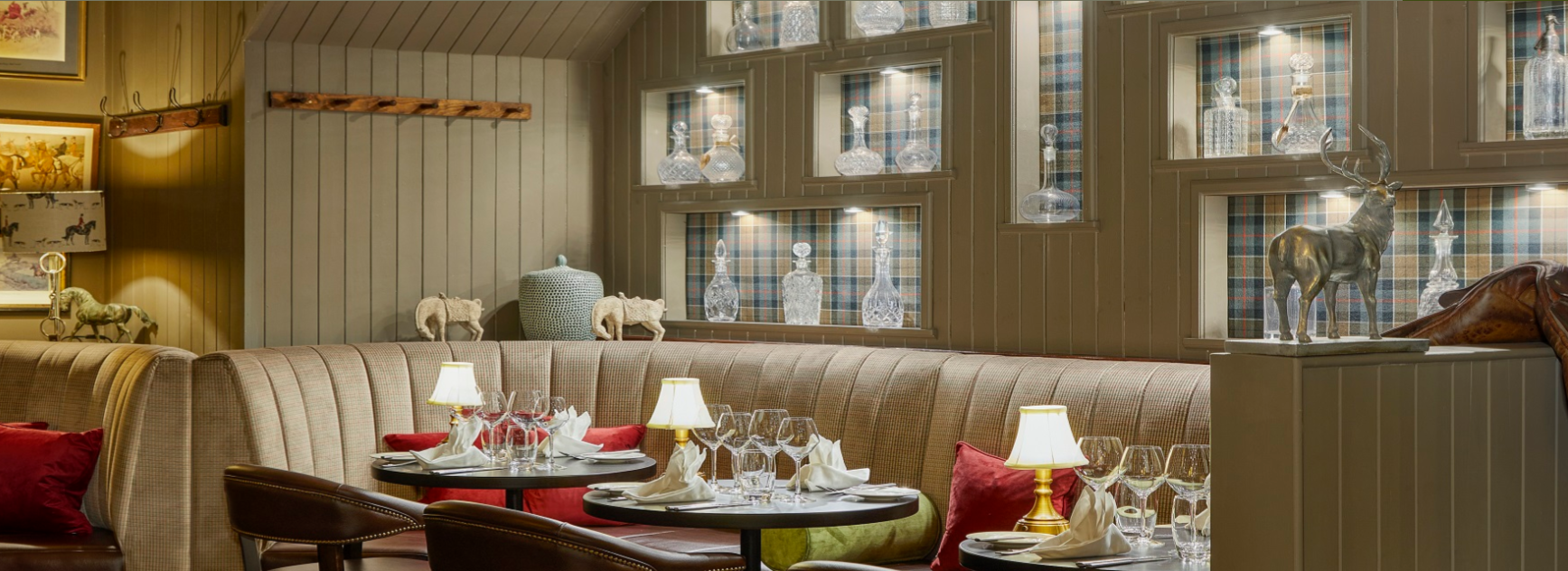 A cozy restaurant interior with a beige upholstered booth, tables set with wine glasses and napkins, small lamps, and decorative items like a metal deer sculpture, glass bottles, and animal figurines on shelves and ledges. The wall has wooden panelin