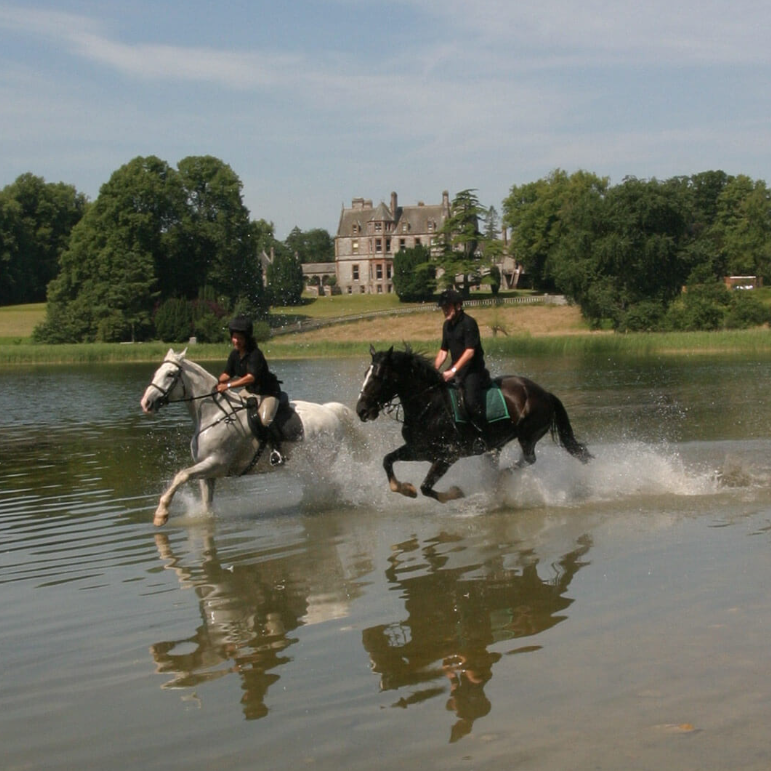 Two riders horseback riding through a shallow body of water, with a large, historic mansion and green trees in the background.
