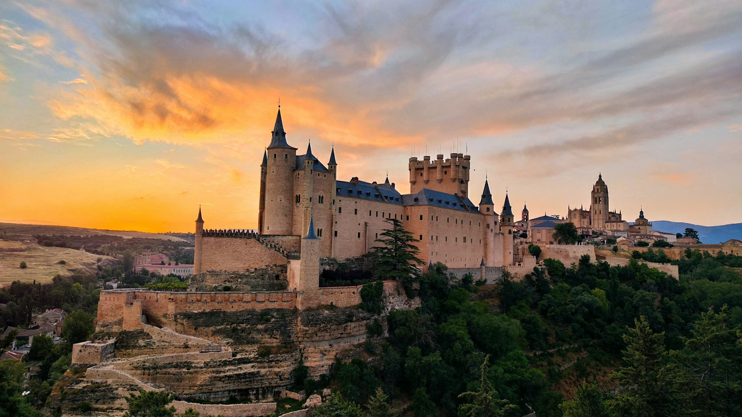 A large medieval castle with towers and walls built on a hill at sunset, overlooking a green landscape with trees and smaller buildings.